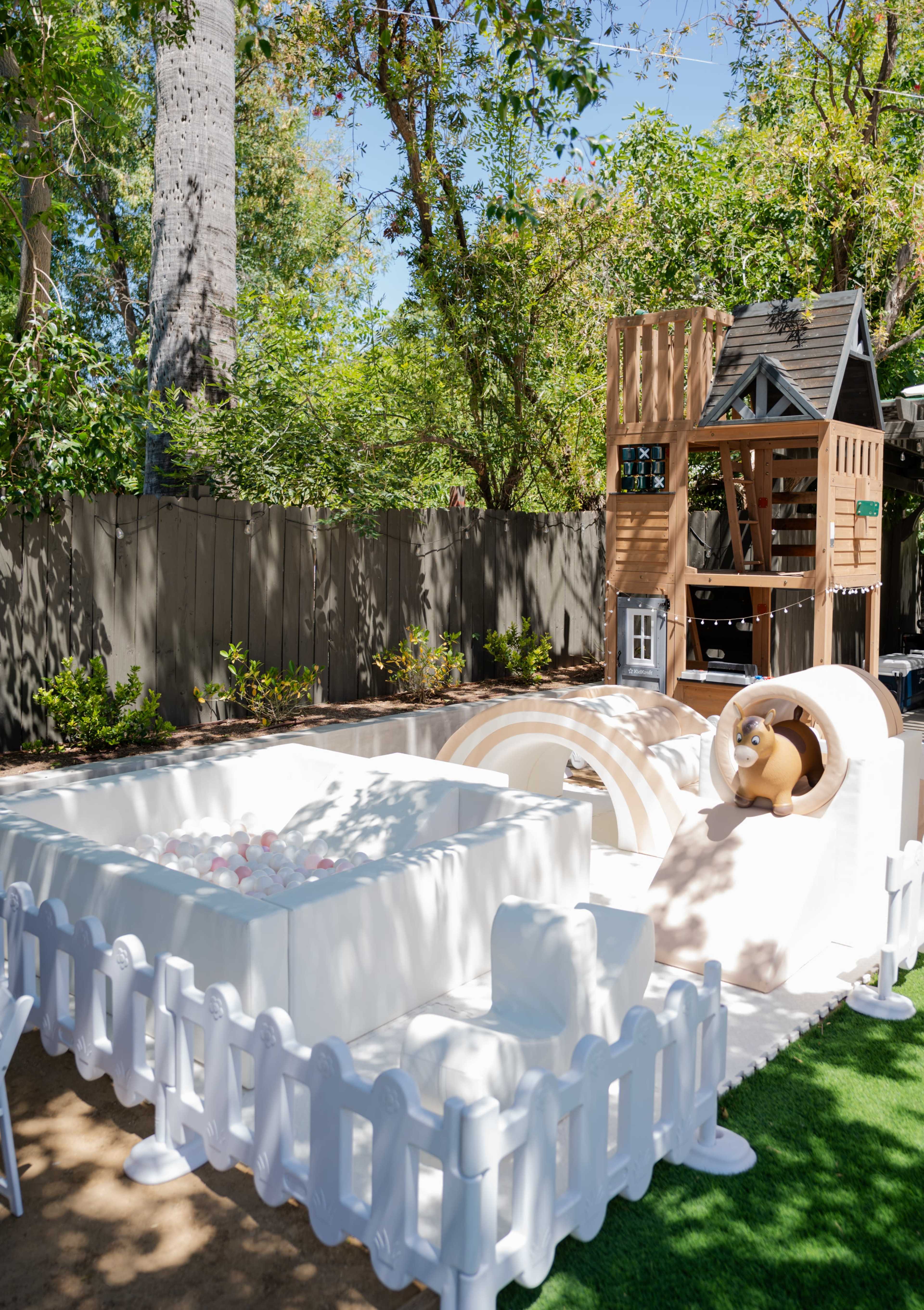 The image shows a children's play area featuring a sandbox, a ball pit, and a raised playhouse surrounded by a white picket fence.