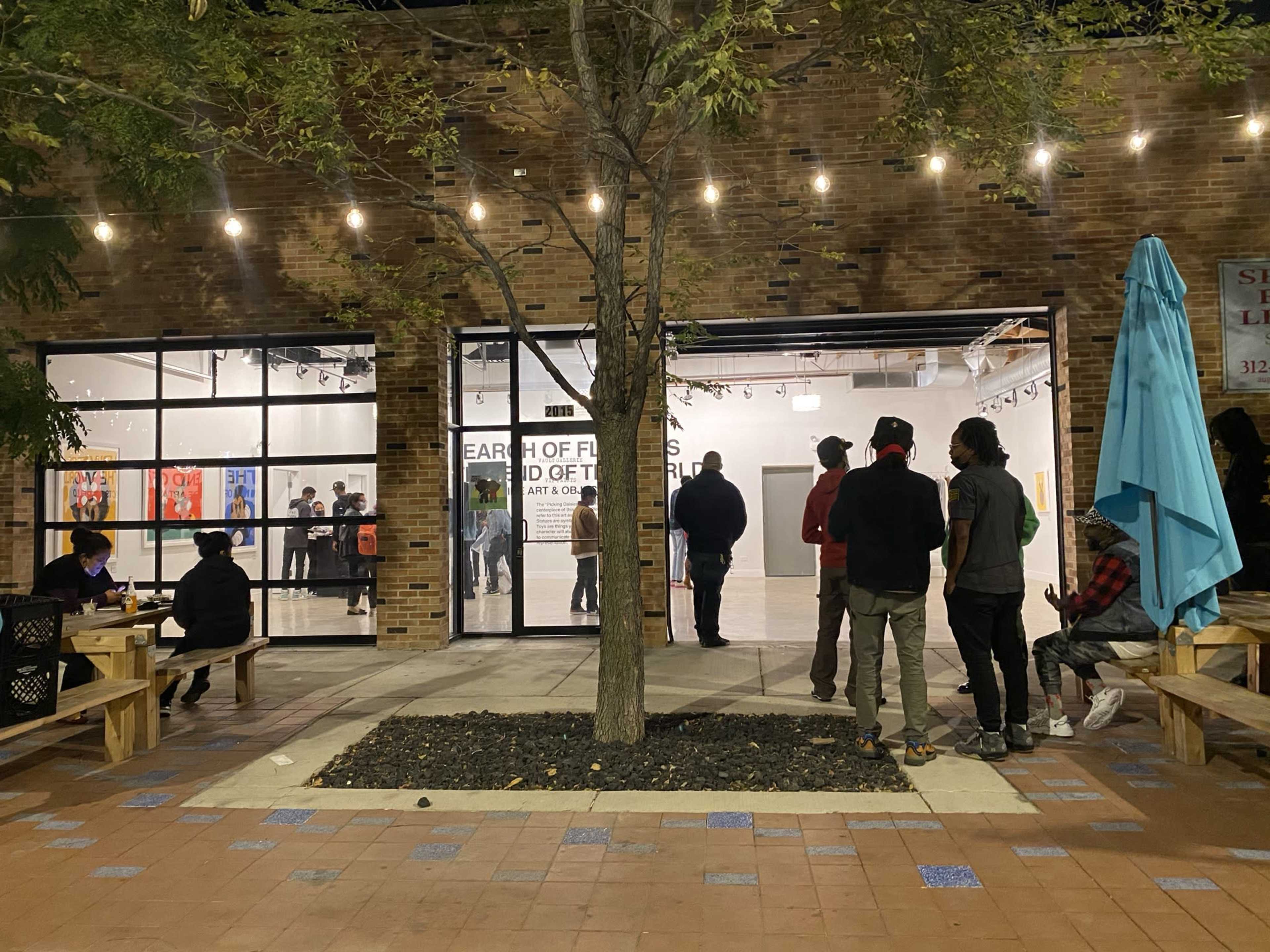 A group of people stands outside an art gallery with illuminated signage and a tree in the center of the courtyard.