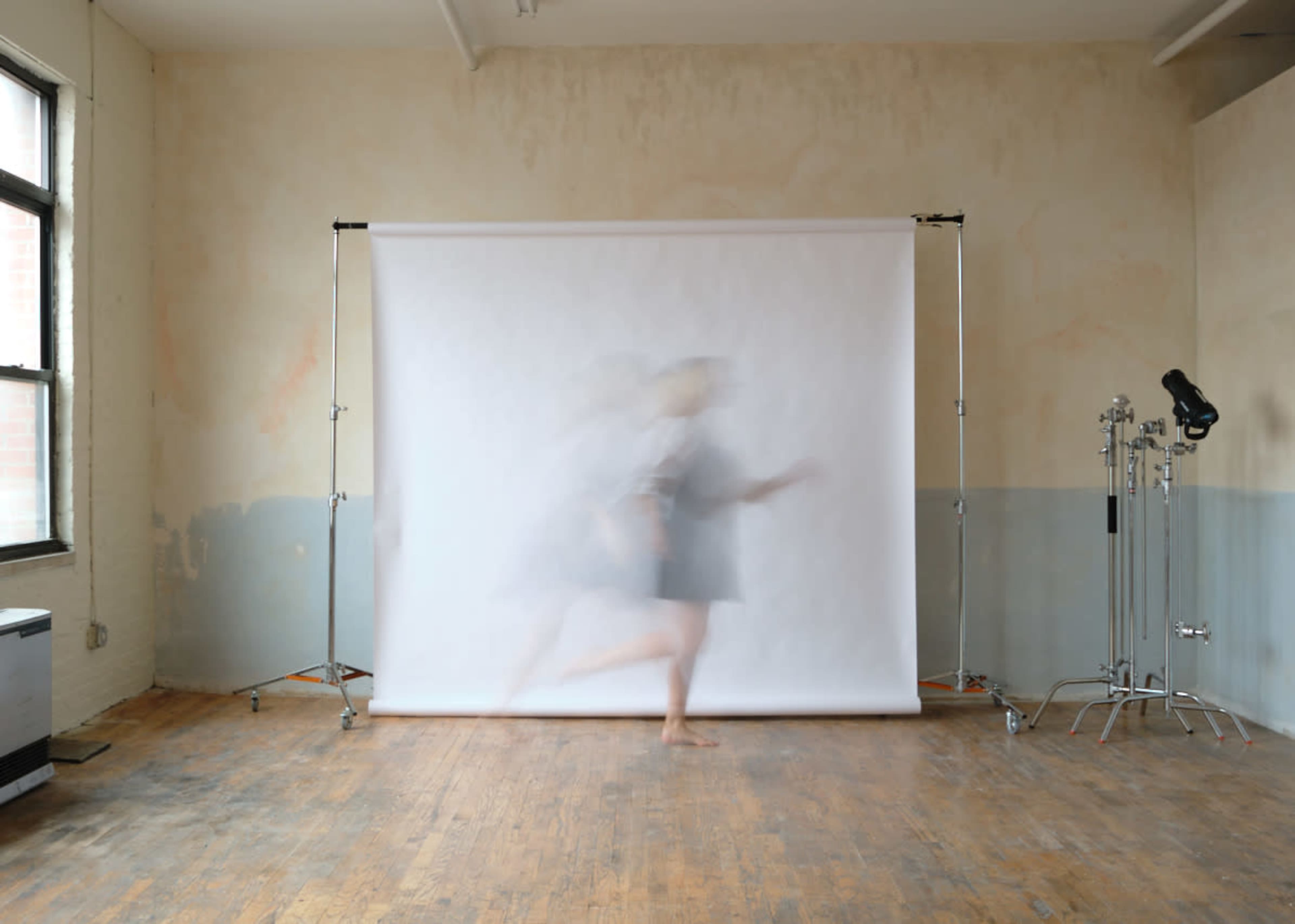 A person runs past a white backdrop in a vacant room with wooden floors and minimal lighting equipment.