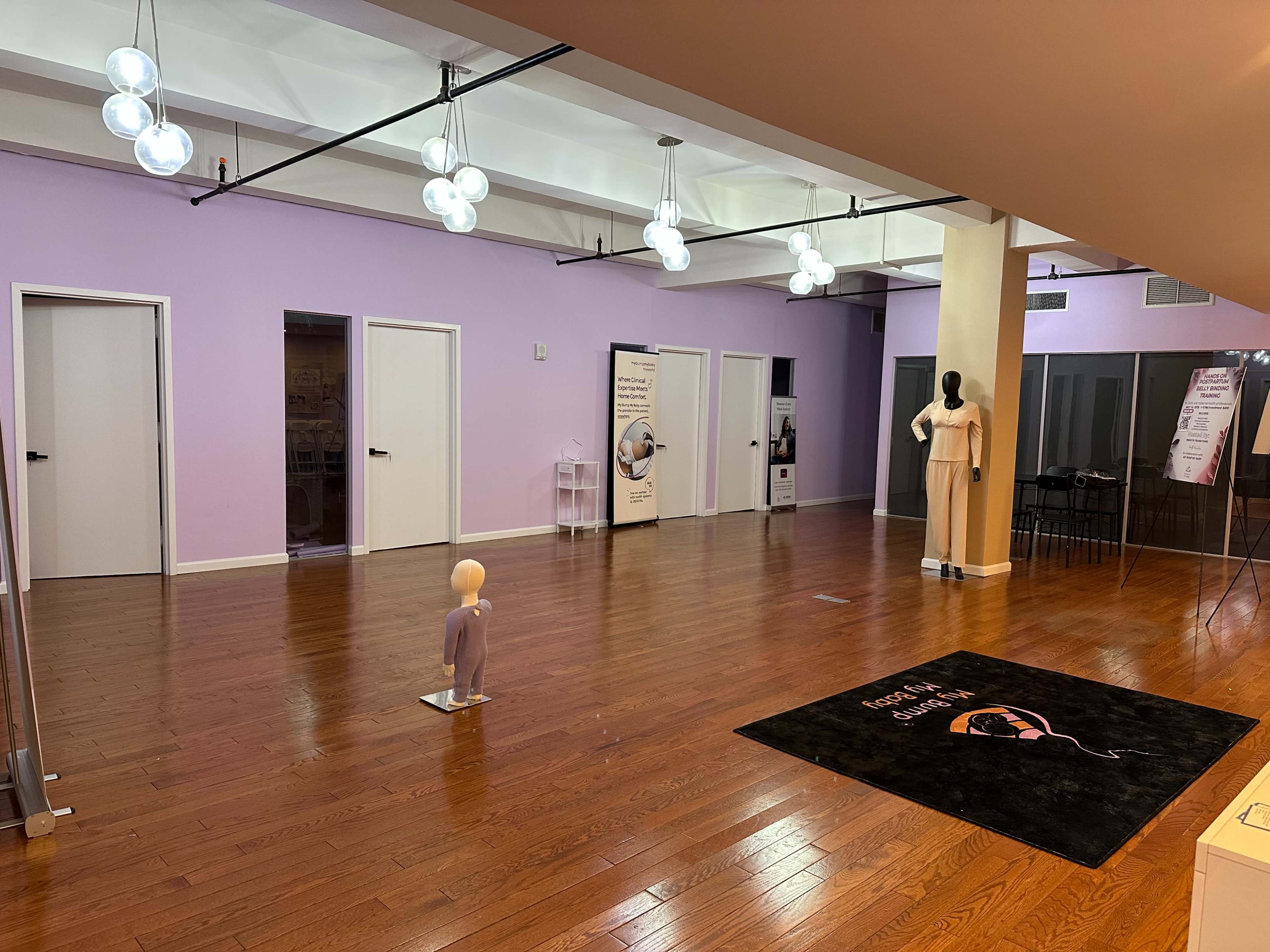 The image shows an empty room with wooden flooring, purple walls, and several closed doors, featuring a mannequin dressed in a light outfit standing on a rug at the entrance.