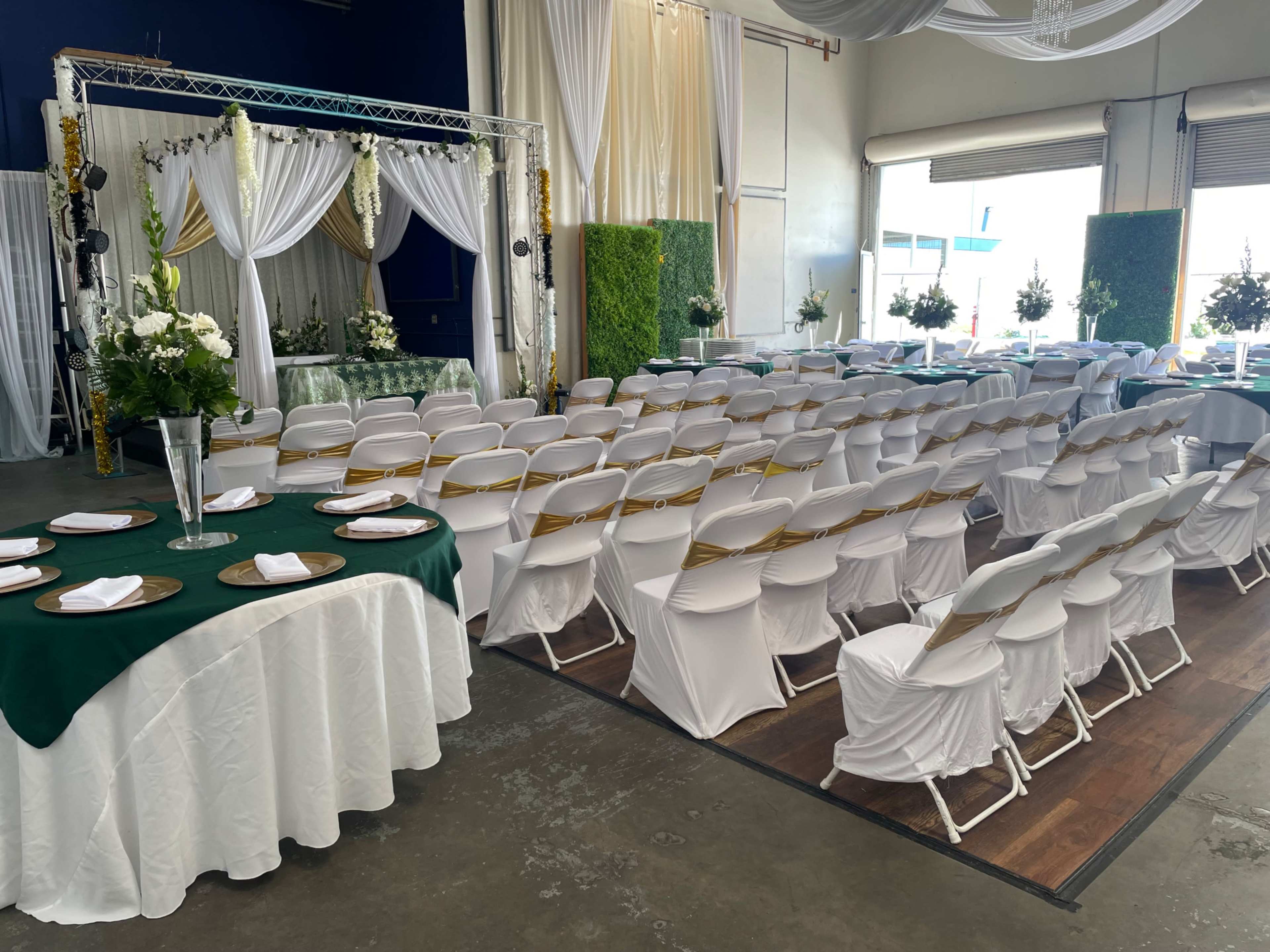 The image shows a decorated indoor venue set up for an event, featuring rows of chairs facing a staged area with elegant fabric draping and floral arrangements.
