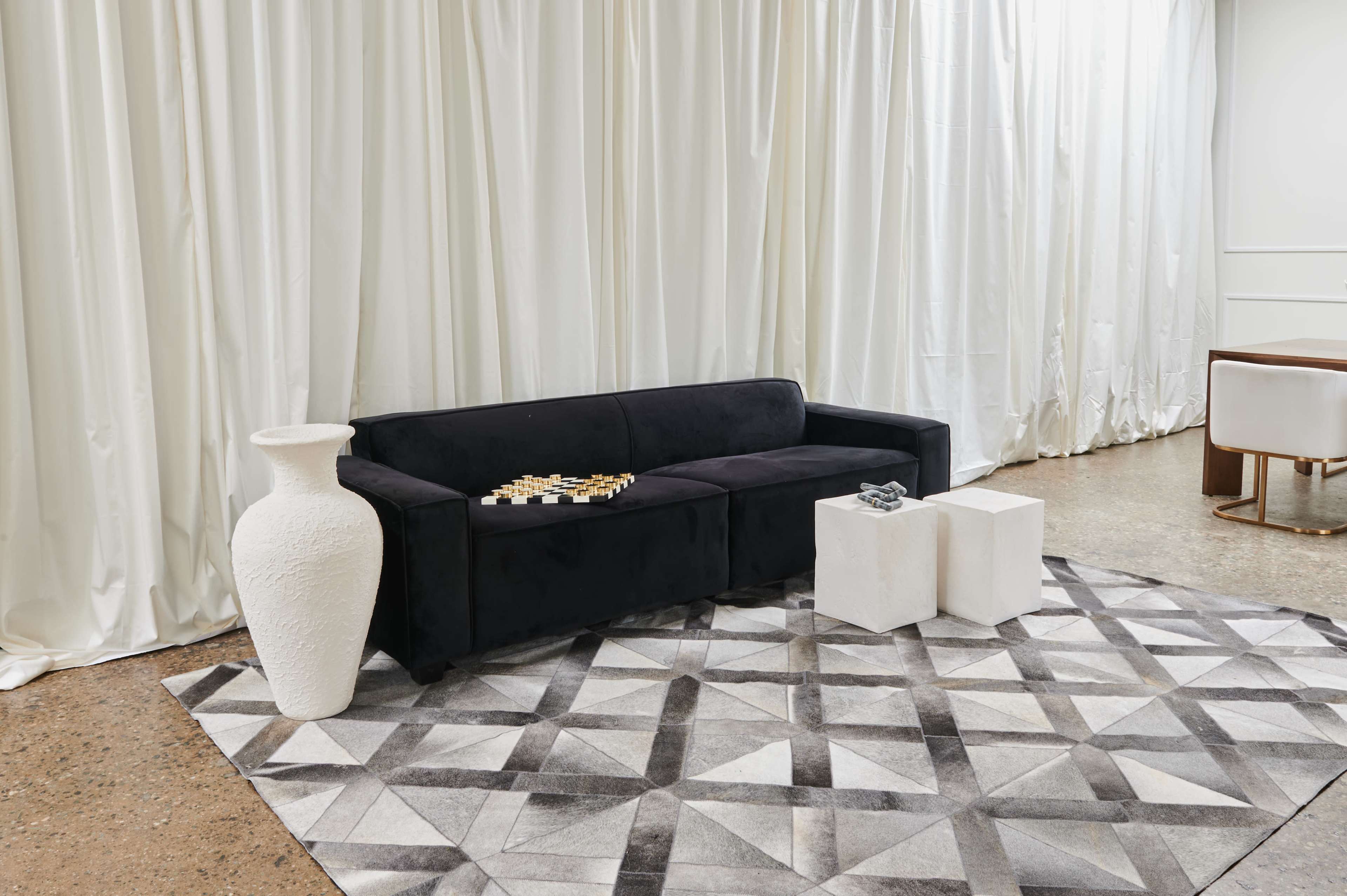 A black sofa with decorative items on it, a white vase, and two white cube tables on a patterned rug in an interior setting.