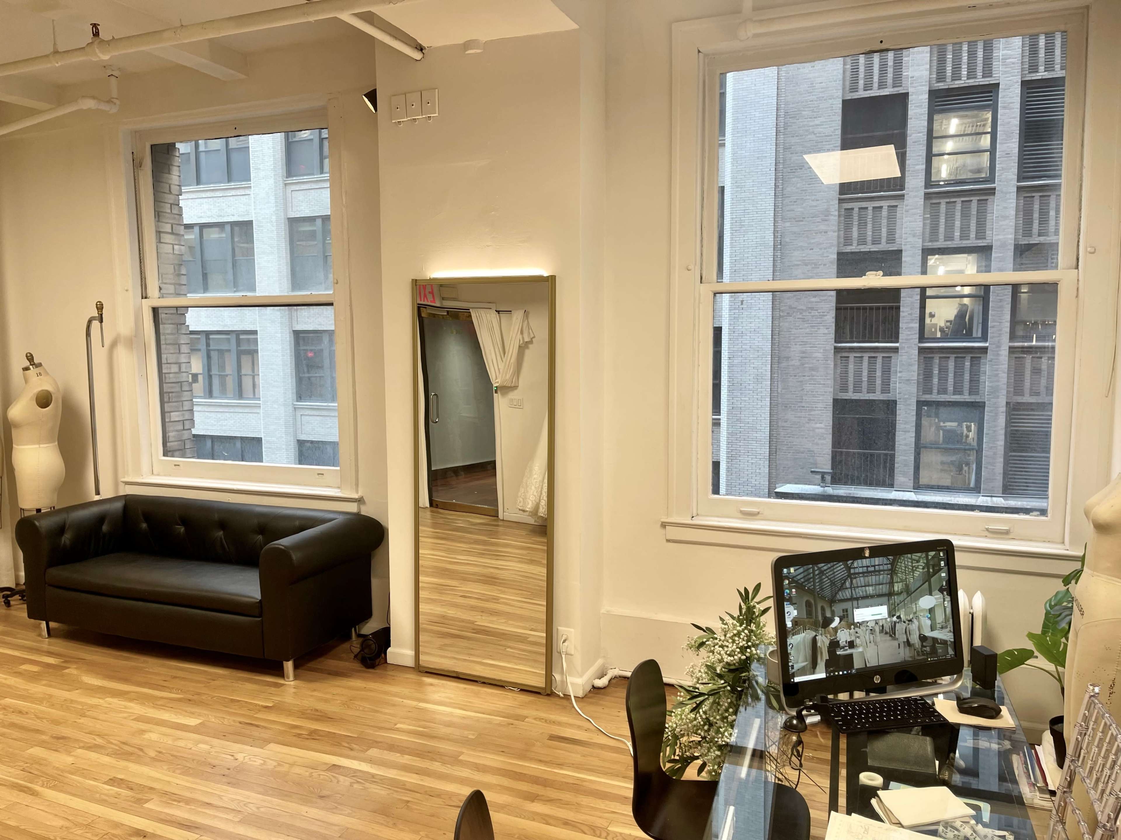 The image shows a minimalist interior of a studio with a black couch, a large mirror, and a desk with a computer near two windows overlooking a building facade.