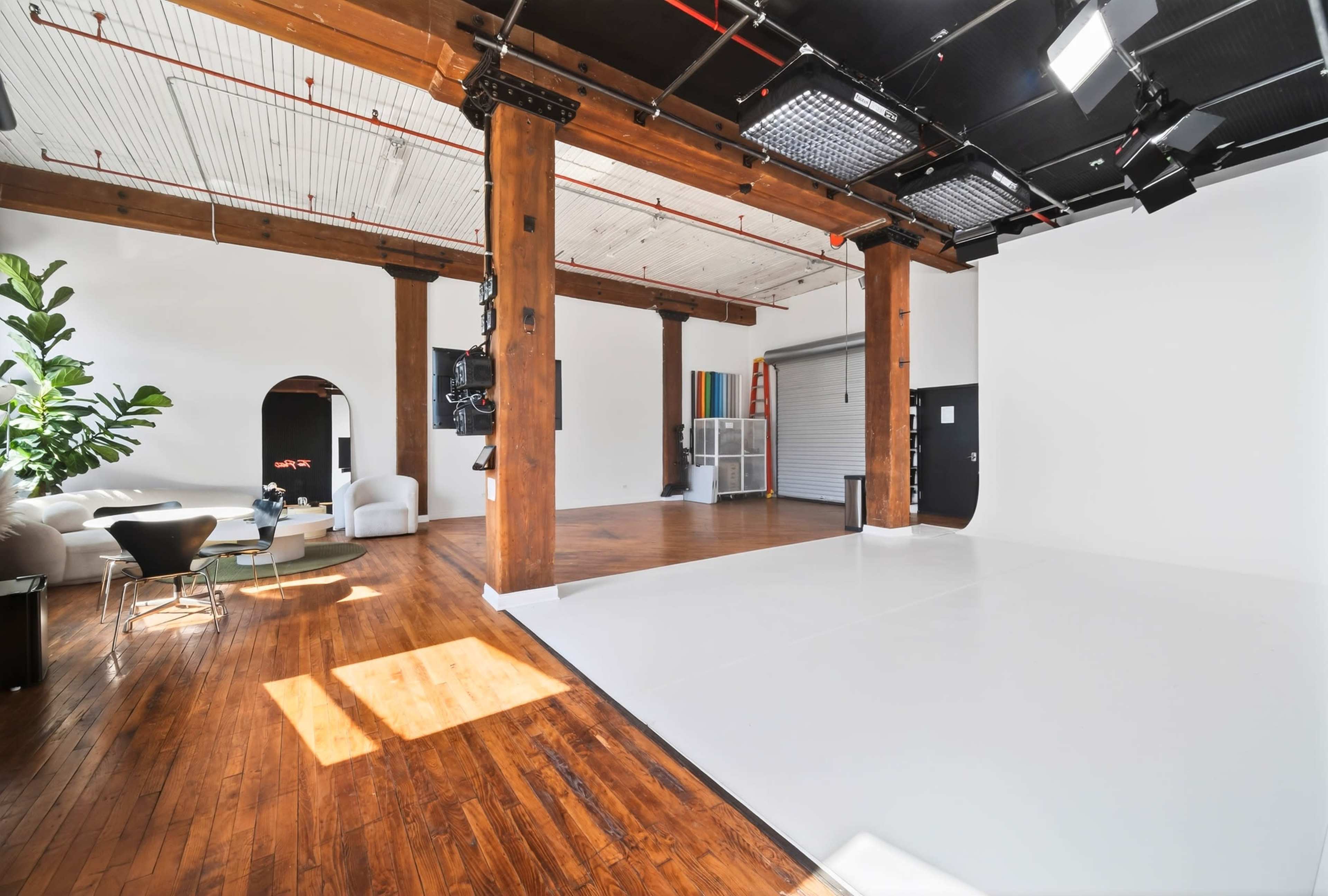 The image shows a spacious, well-lit studio with wooden beams, polished hardwood floors, and a white backdrop area.