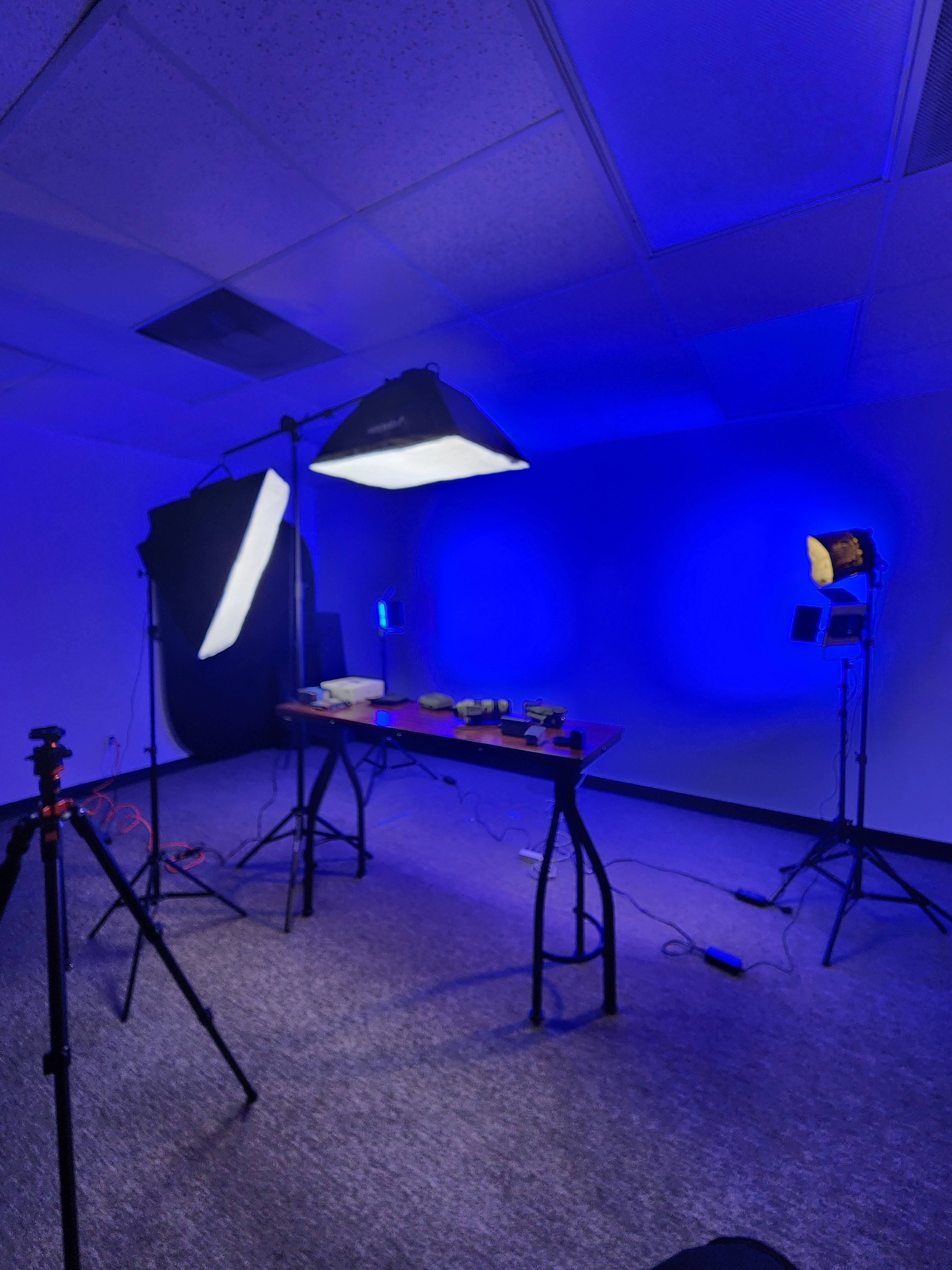 The image shows a dimly lit studio setup with blue lighting, featuring a table and various equipment, including light stands and a backdrop.