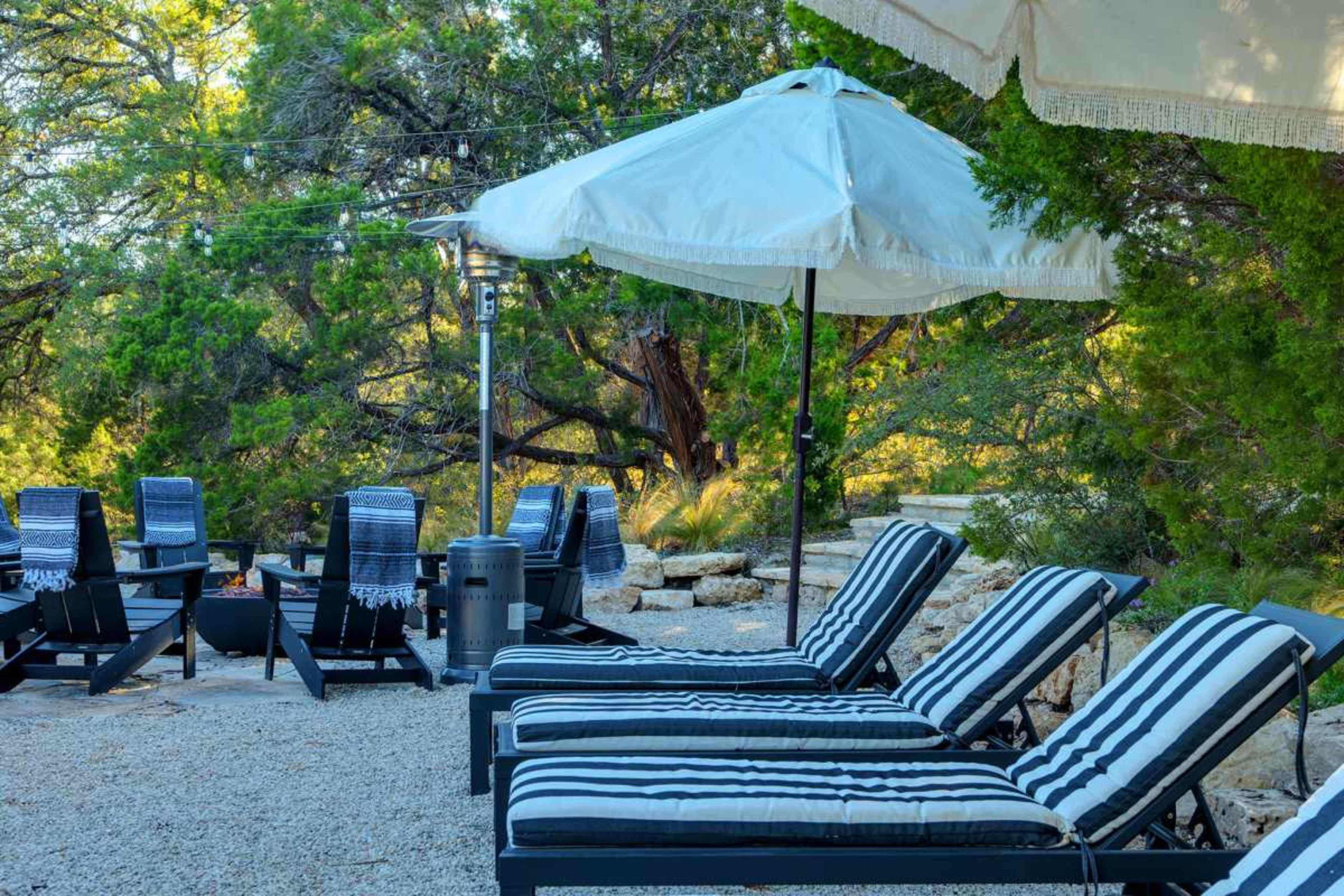 A patio area features black and white striped lounge chairs, an umbrella, and a seating arrangement around a fire pit amidst trees and gravel.