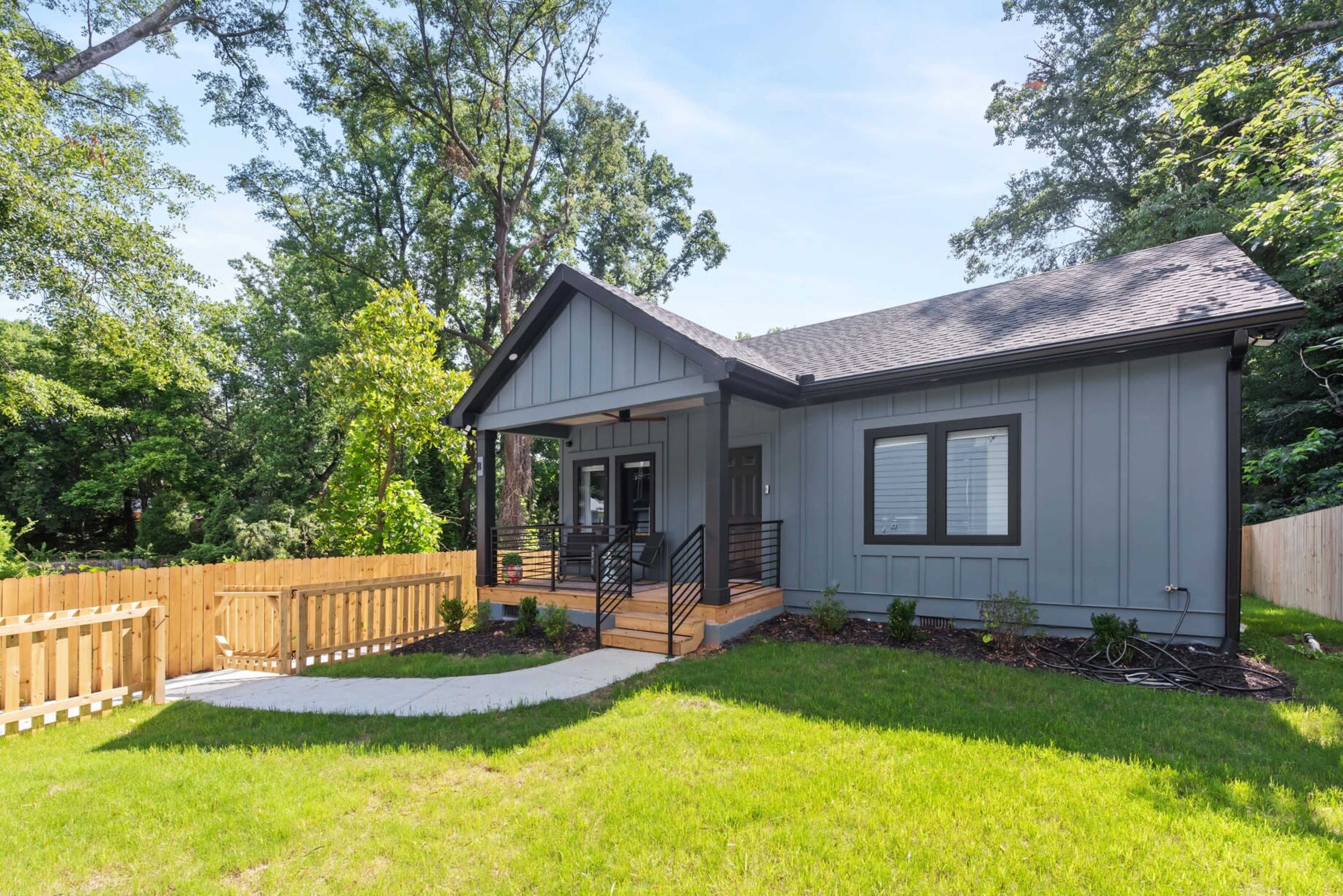 The image shows a contemporary single-story house with a gray exterior, a wood front porch, and a landscaped yard surrounded by a wooden fence.
