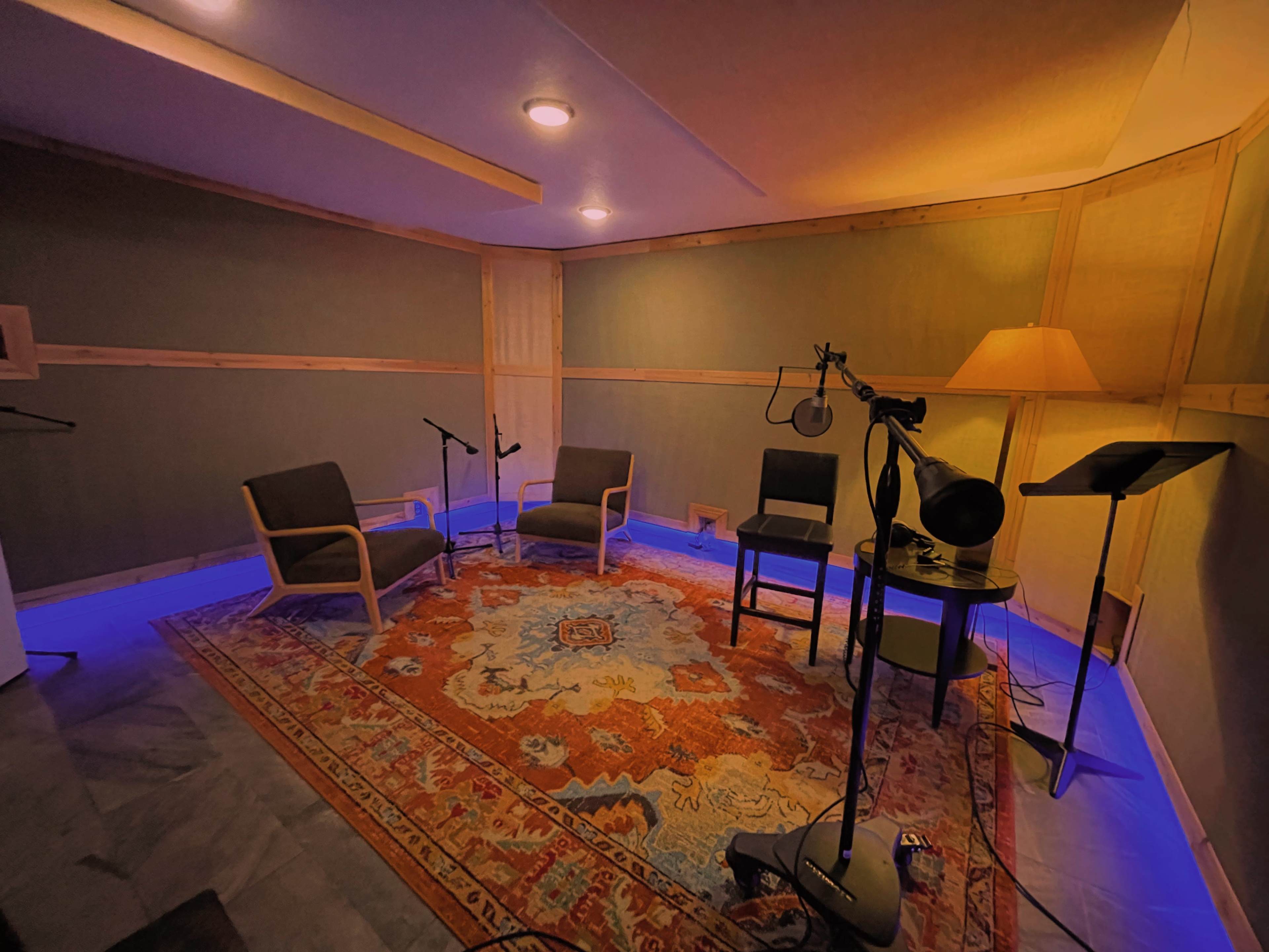 A soundproof recording studio with two chairs, a microphone on a stand, and a floor lamp, illuminated by blue lighting.