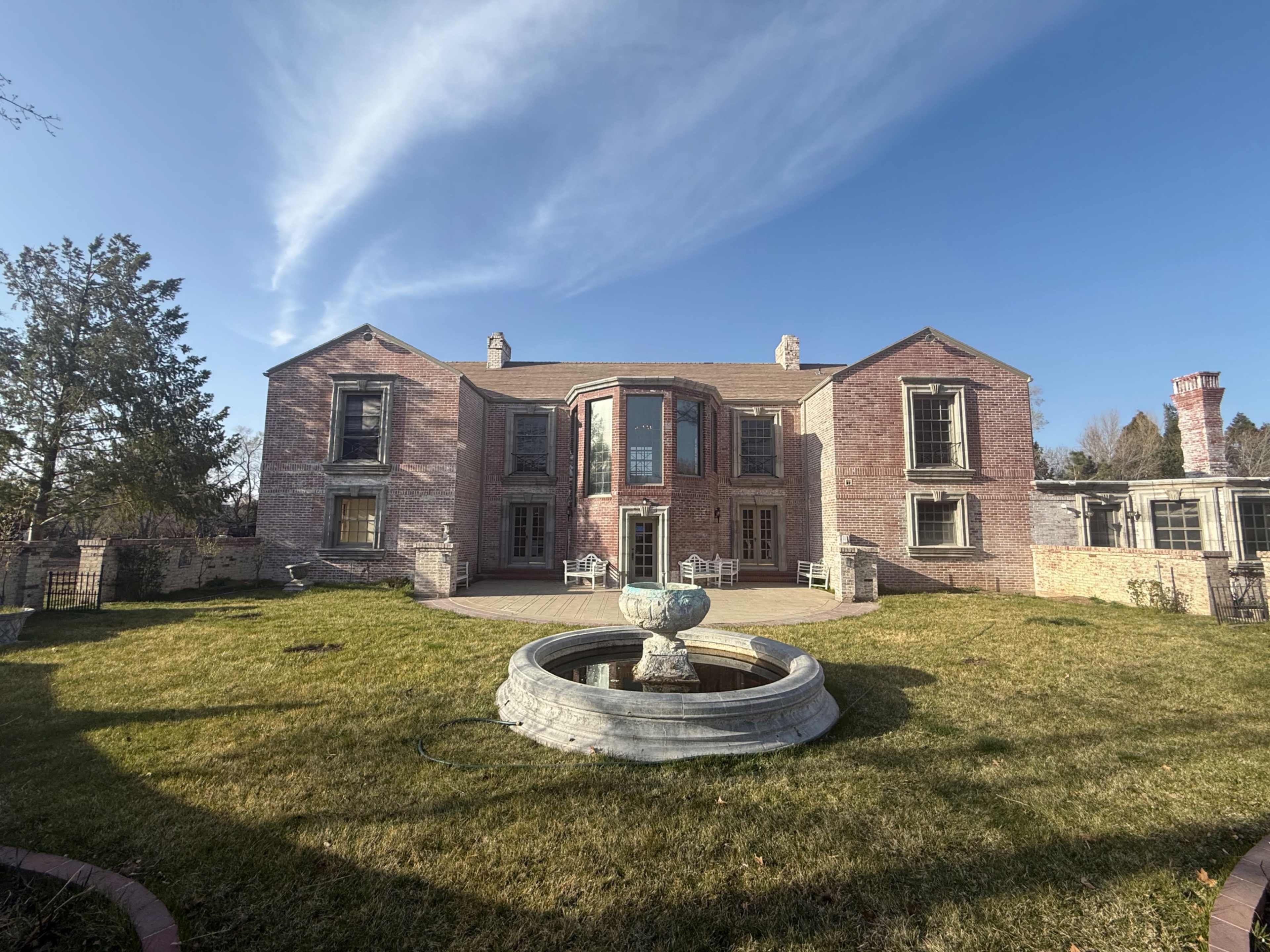 The image shows a large brick mansion with a central circular bay, surrounded by a grassy lawn and a circular fountain in the foreground.