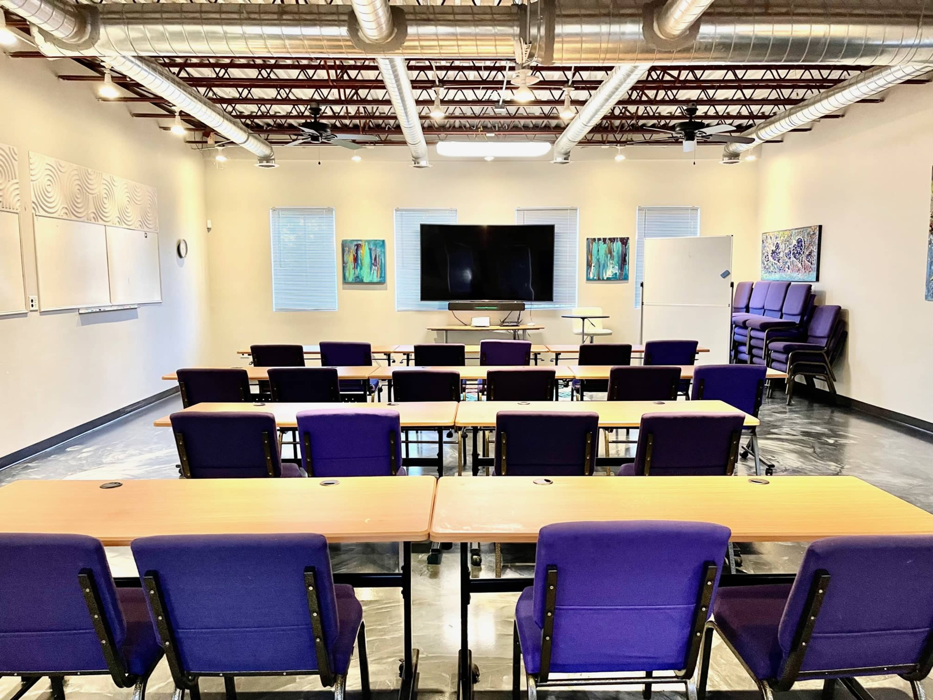 The image shows a modern classroom with rows of purple chairs and tables facing a large screen and whiteboard in a well-lit space.