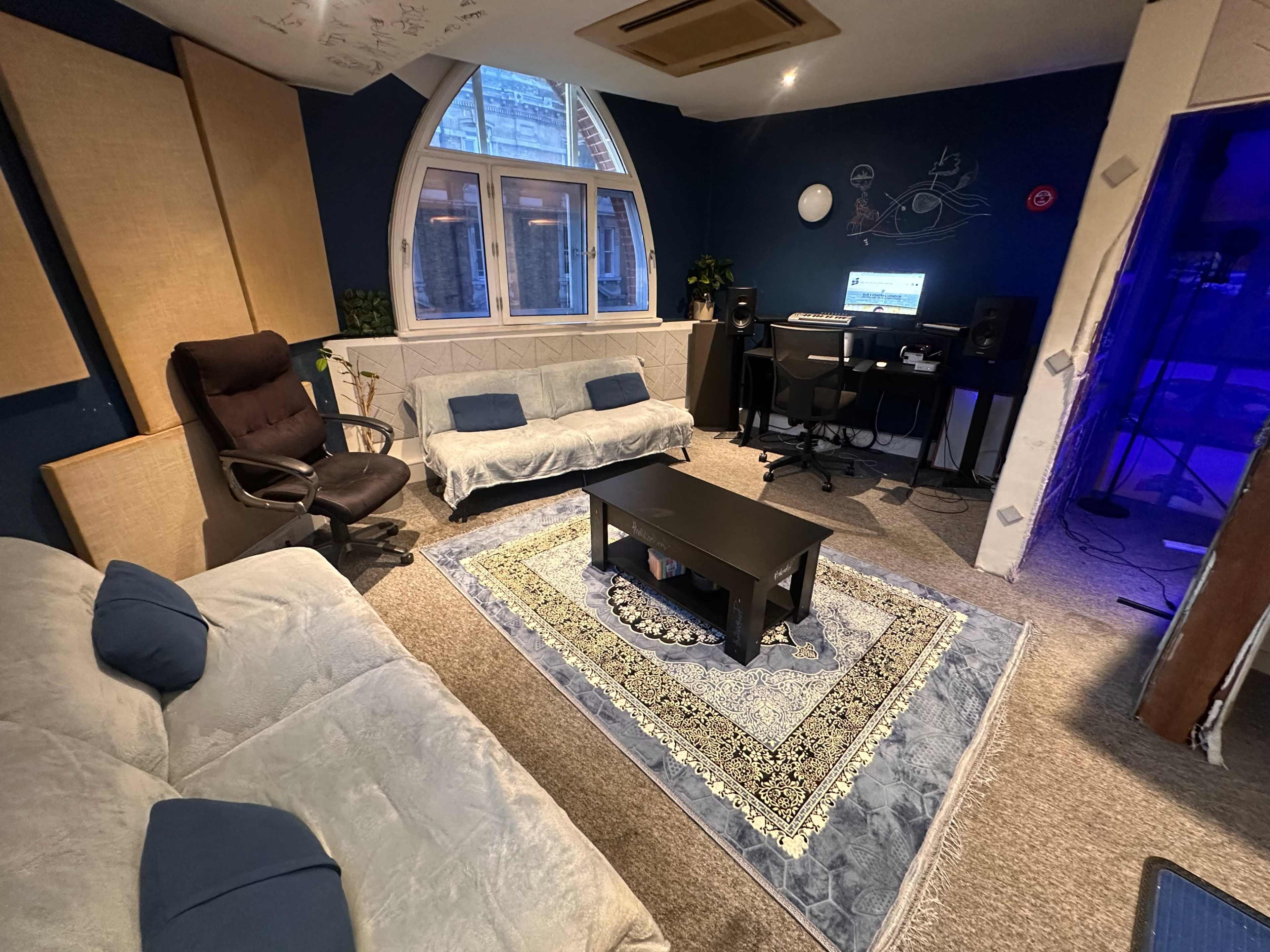 The image shows a cozy studio-like room with two sofas, a coffee table, a desk with computer equipment, and large windows.