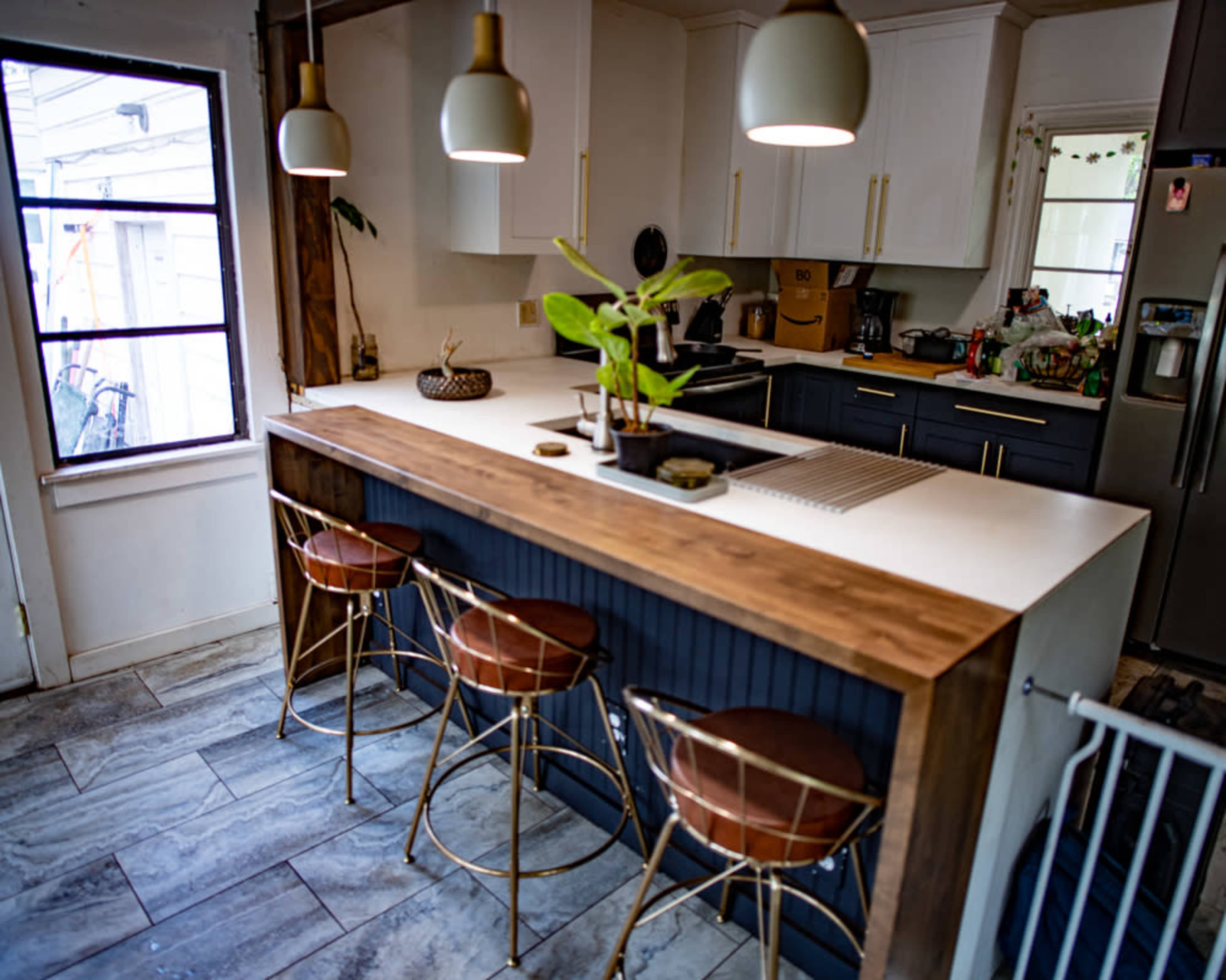 The image shows a modern kitchen with a central island featuring three bar stools, surrounded by cabinets and appliances.