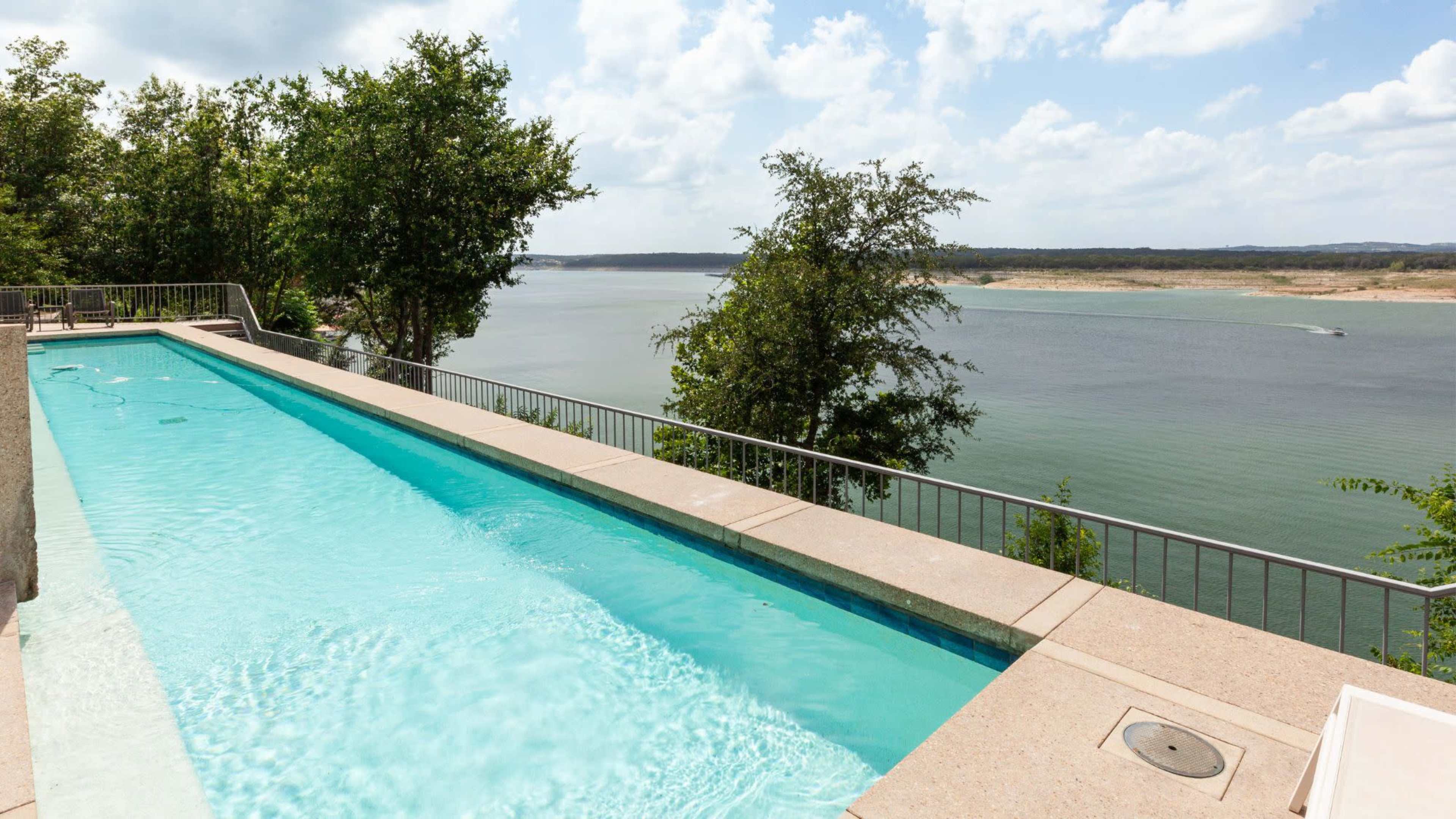 A long, narrow swimming pool is situated on a hillside overlooking a body of water, with trees providing some shade along the edge.