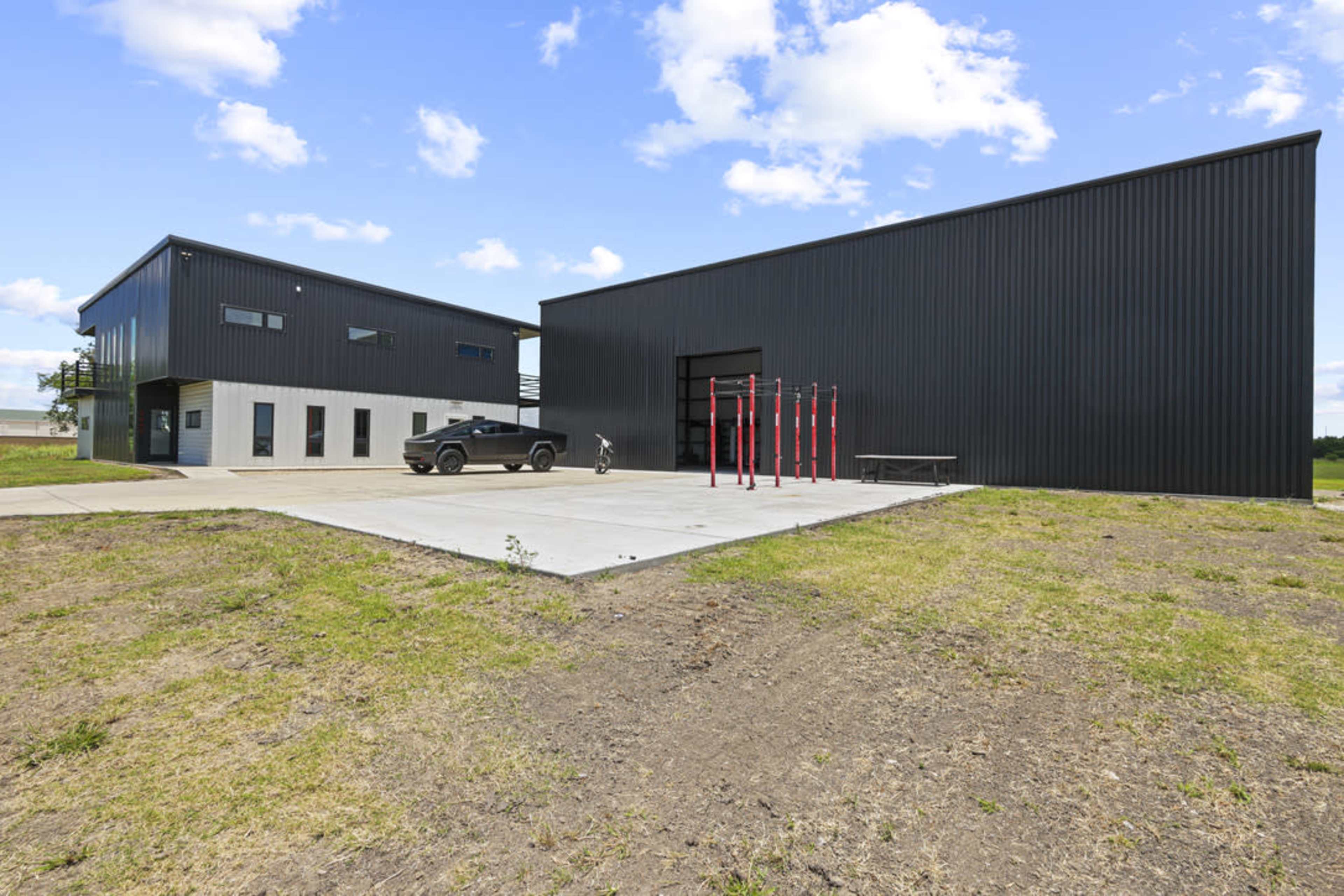 The image shows a modern, black metal building complex situated on a grassy lot with a parked vehicle and outdoor exercise equipment.