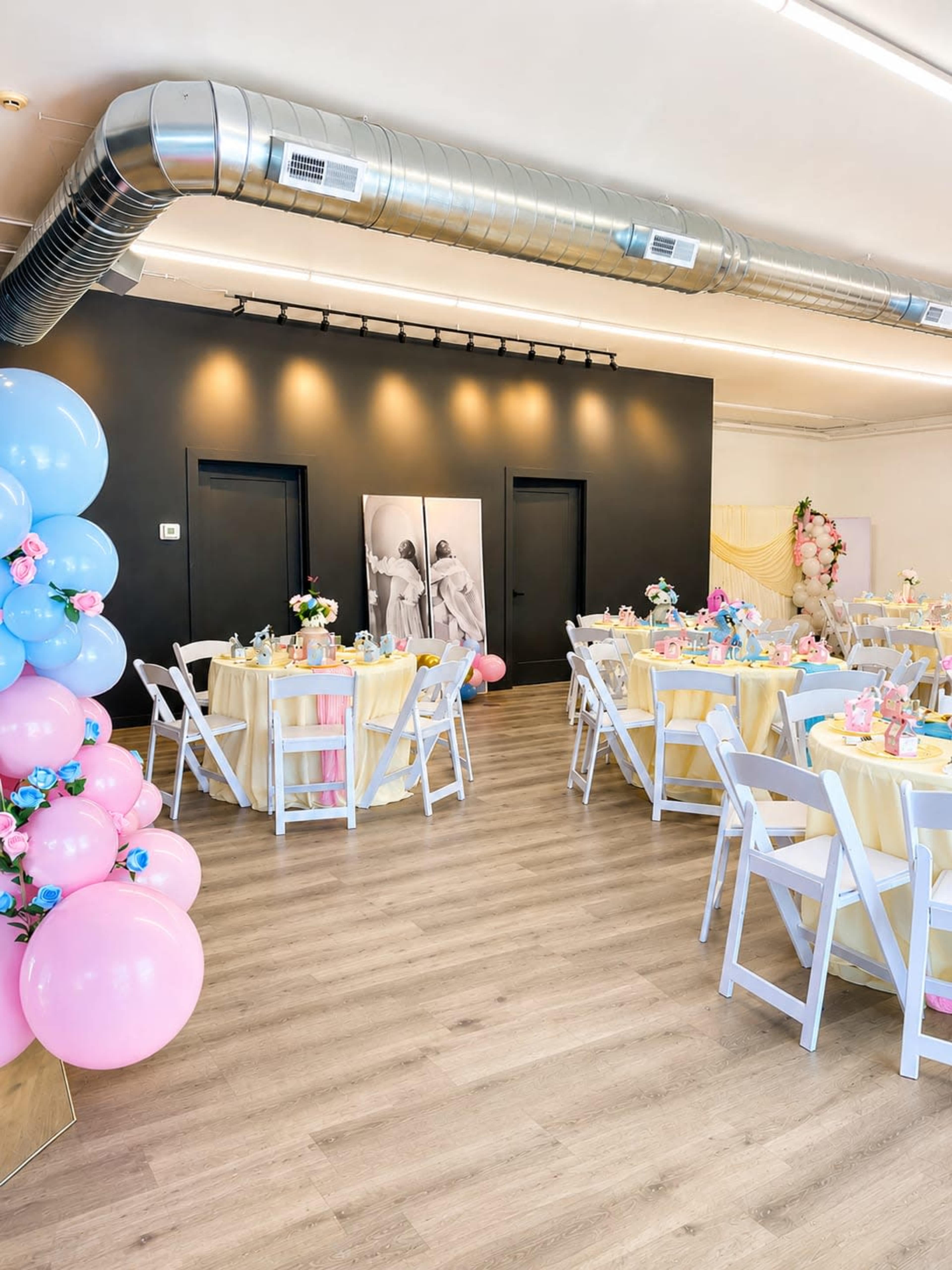 The image shows a decorated event space featuring round tables with yellow tablecloths, colorful centerpieces, and an arrangement of balloon decorations in blue and pink along one side.
