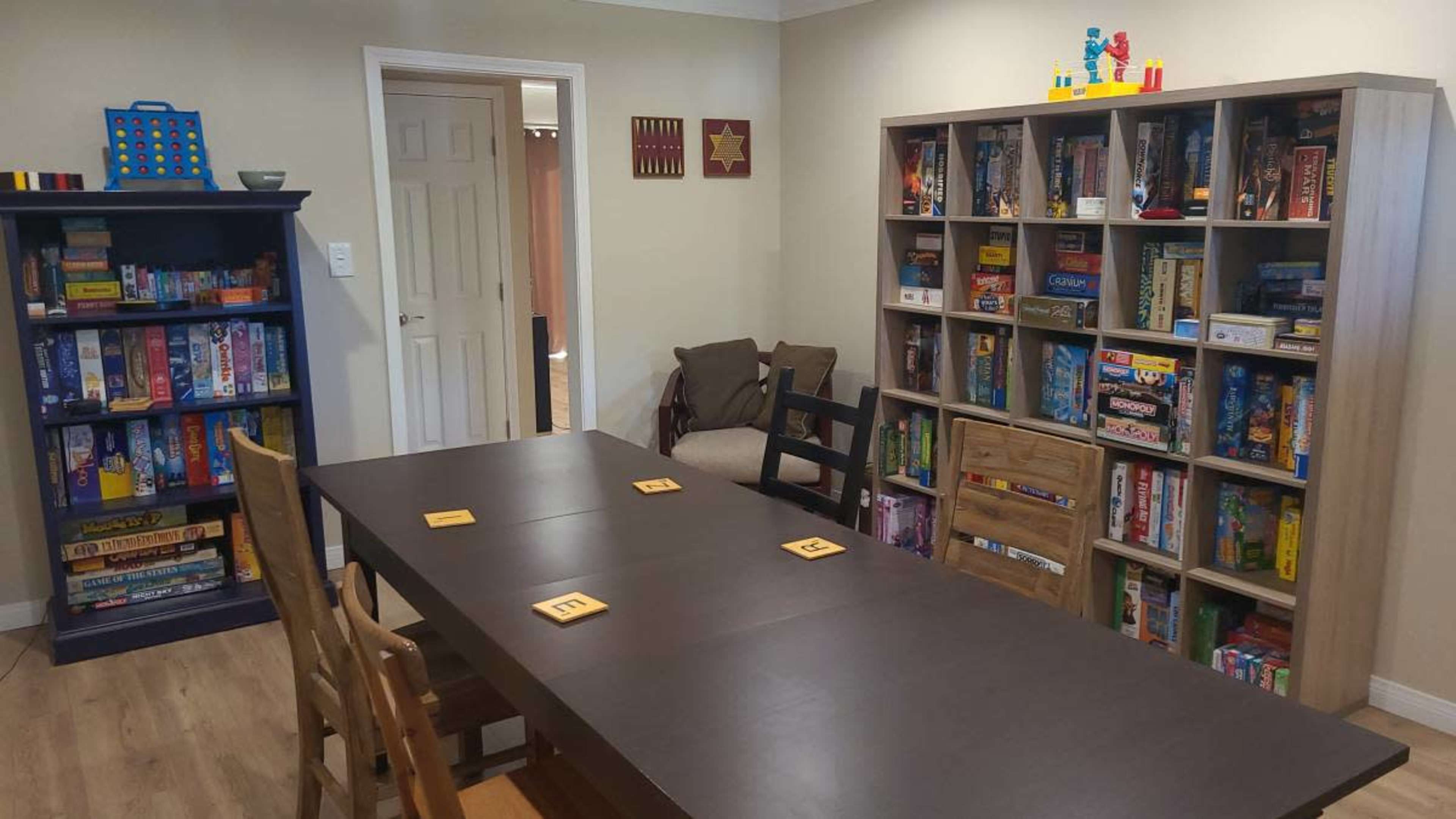 A game room features a large table surrounded by chairs, with shelves filled with various board games on either side.