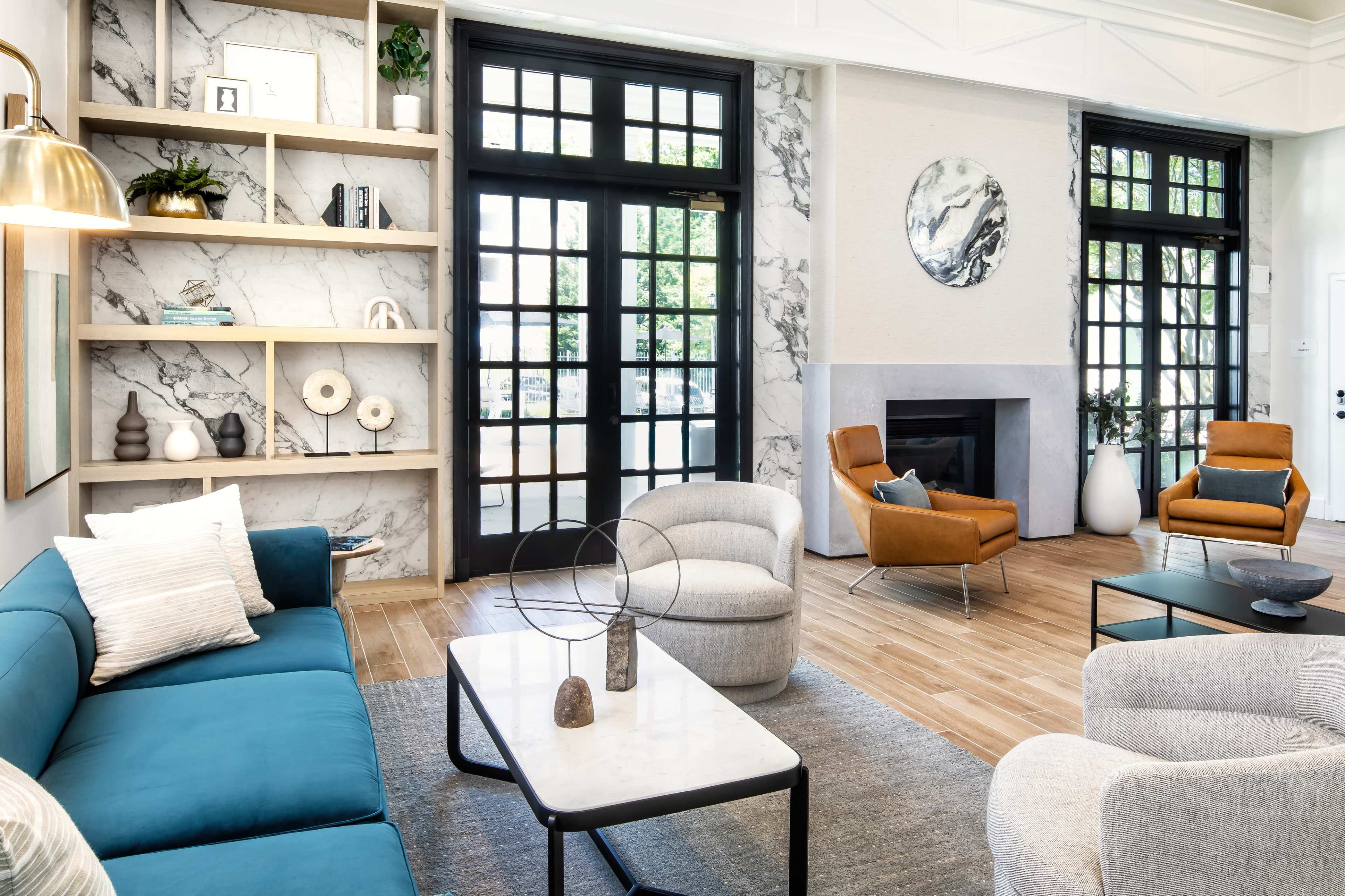 The image shows a modern living room featuring a blue sofa, a marble coffee table, and two accent chairs, with large windows allowing natural light to fill the space.