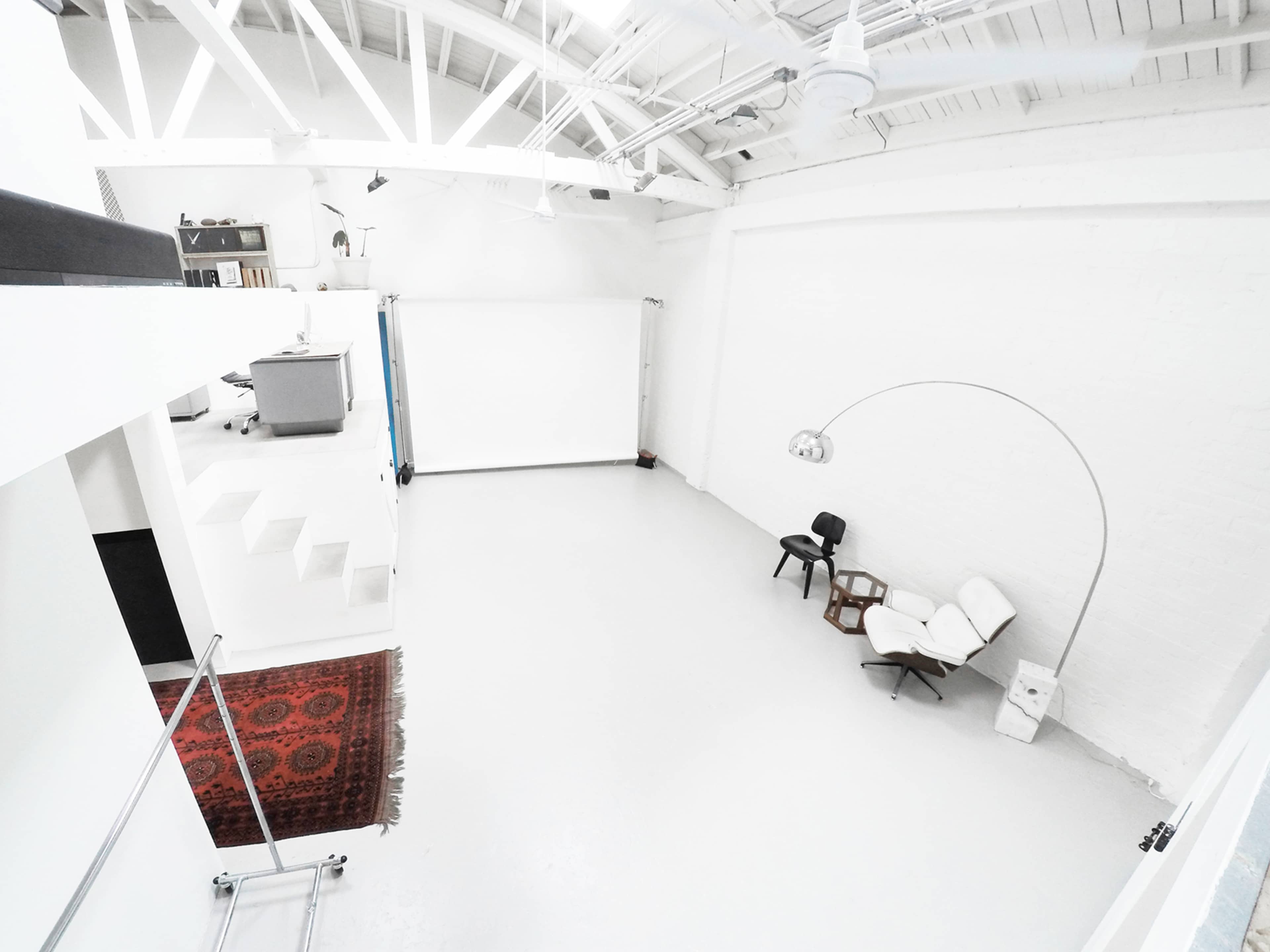 A spacious, industrial-style room with white walls, a light gray floor, minimal furniture, and a large white screen.