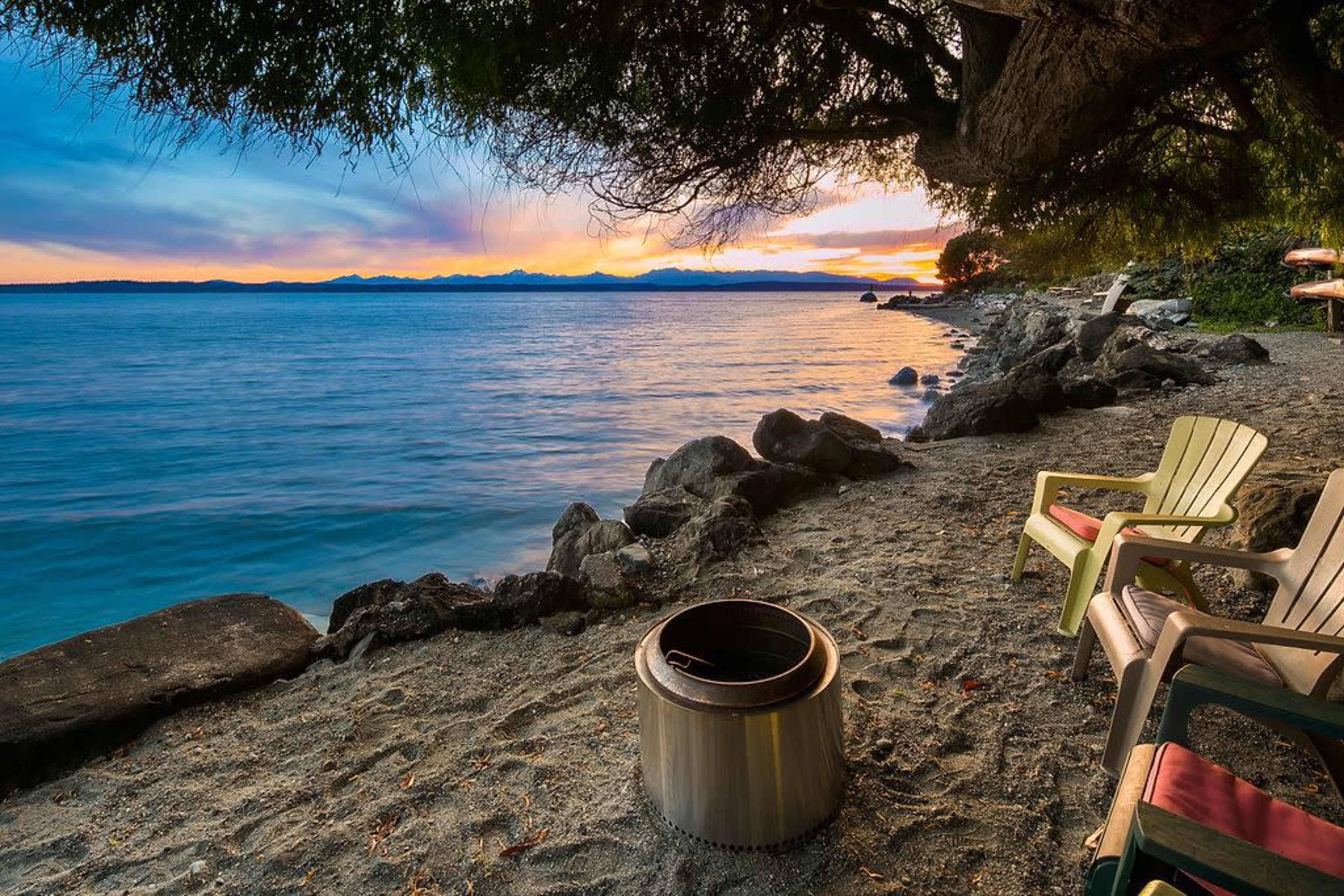 A peaceful beach scene at sunset, with a fire pit surrounded by adirondack chairs on the sandy shore.
