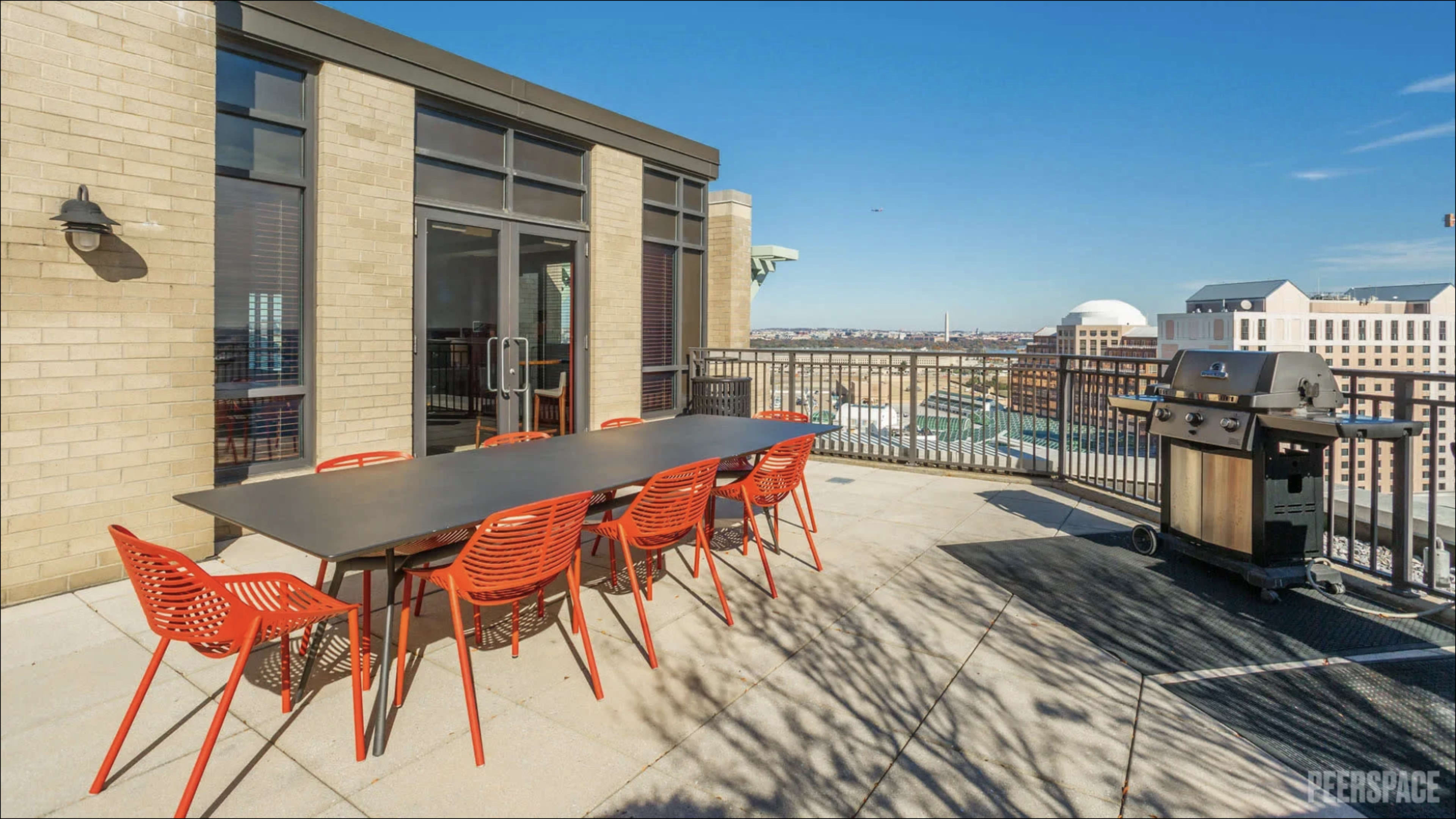 The image shows a rooftop terrace featuring a long black table with red chairs and a grill, overlooking a cityscape under a clear blue sky.