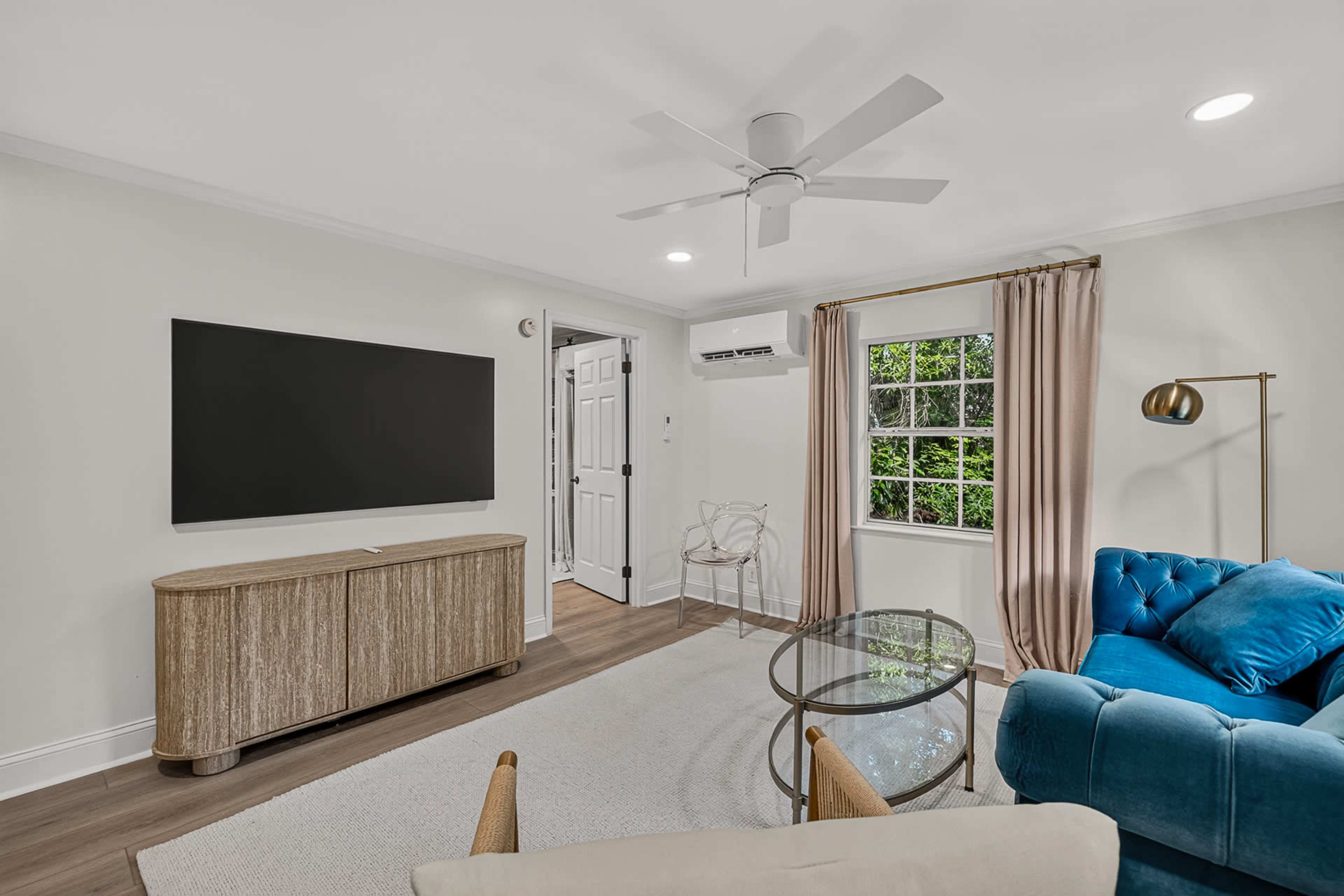 A modern living room features a blue sofa, a glass coffee table, a large television mounted on the wall, and a window with curtains.