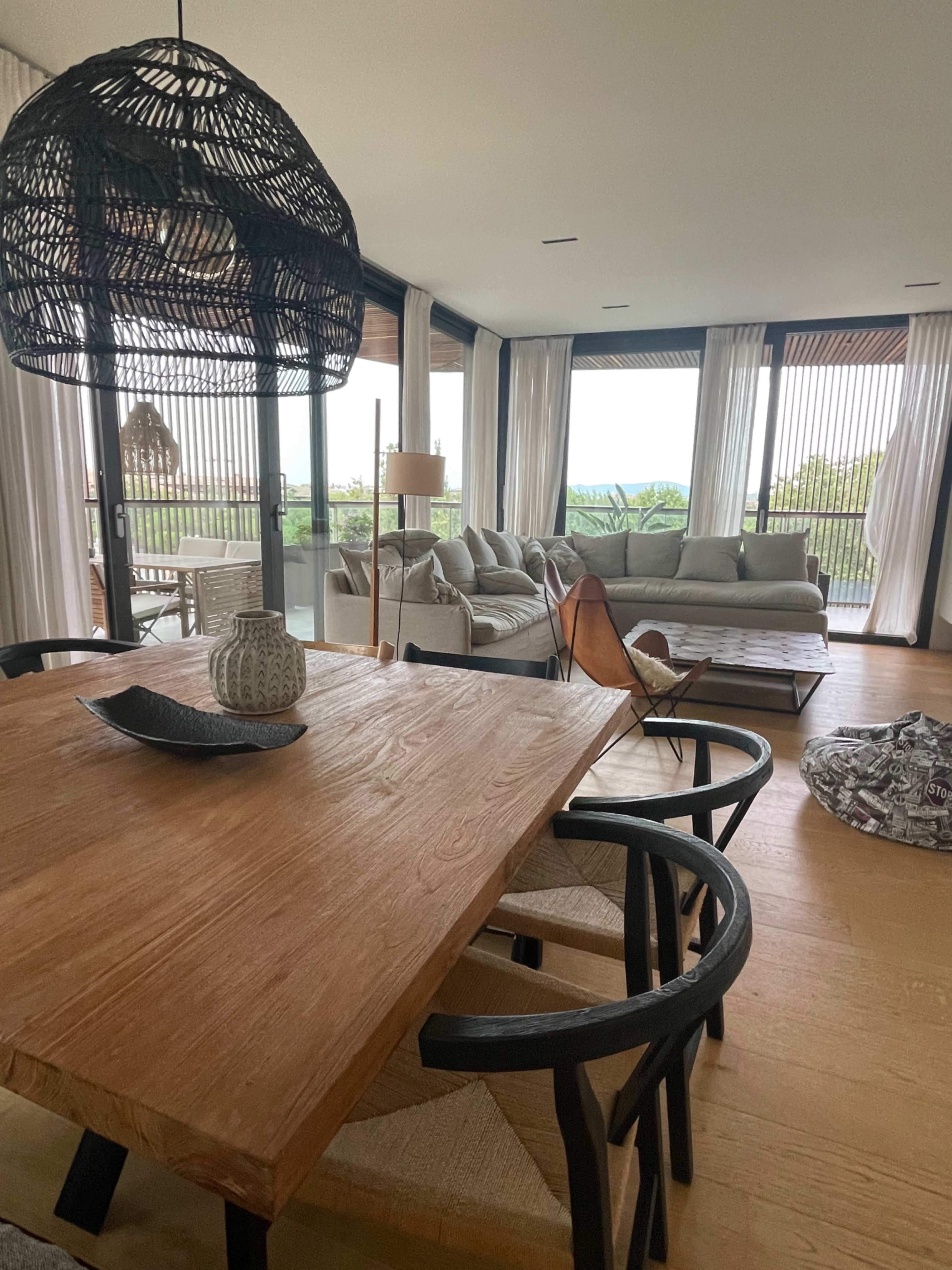 The image depicts a modern living space with a wooden dining table, sleek chairs, and large windows overlooking a green landscape.