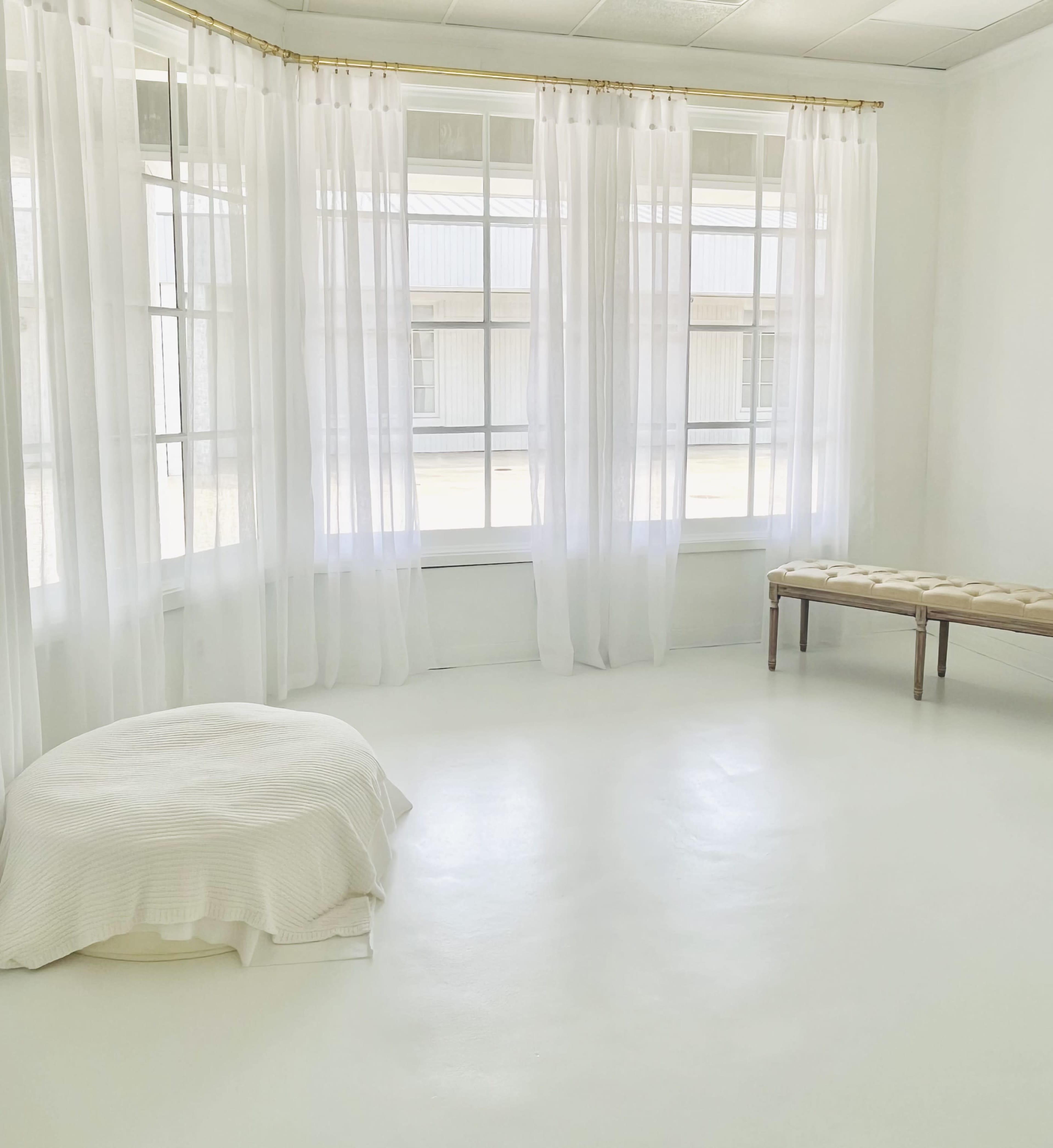 The image shows a bright, minimalistic room with large windows draped in white curtains, a round white cushion on the floor, and a wooden bench against the wall.