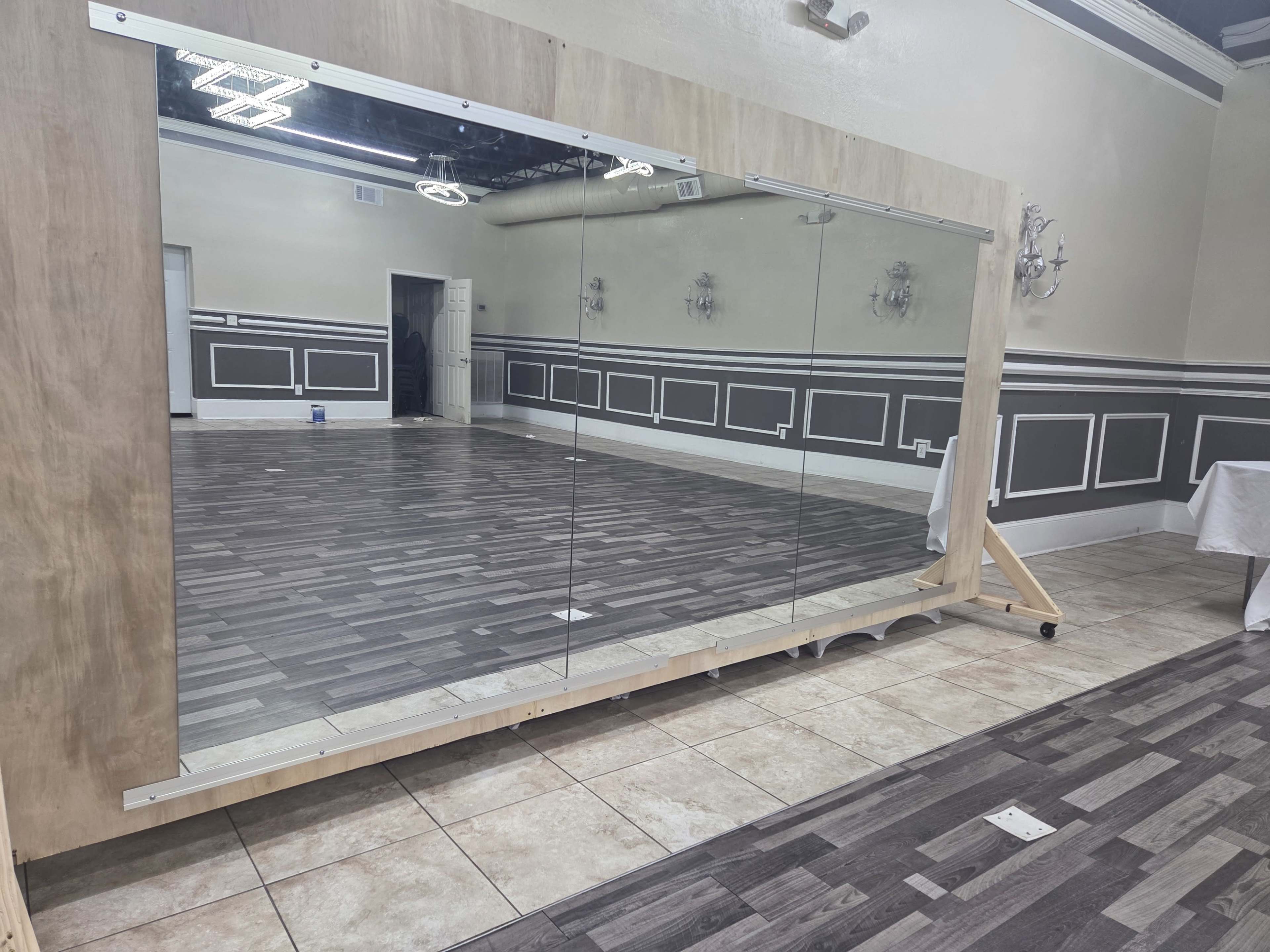 A large, framed mirror is positioned against a wall in a spacious room with wooden flooring and decorative wall paneling.