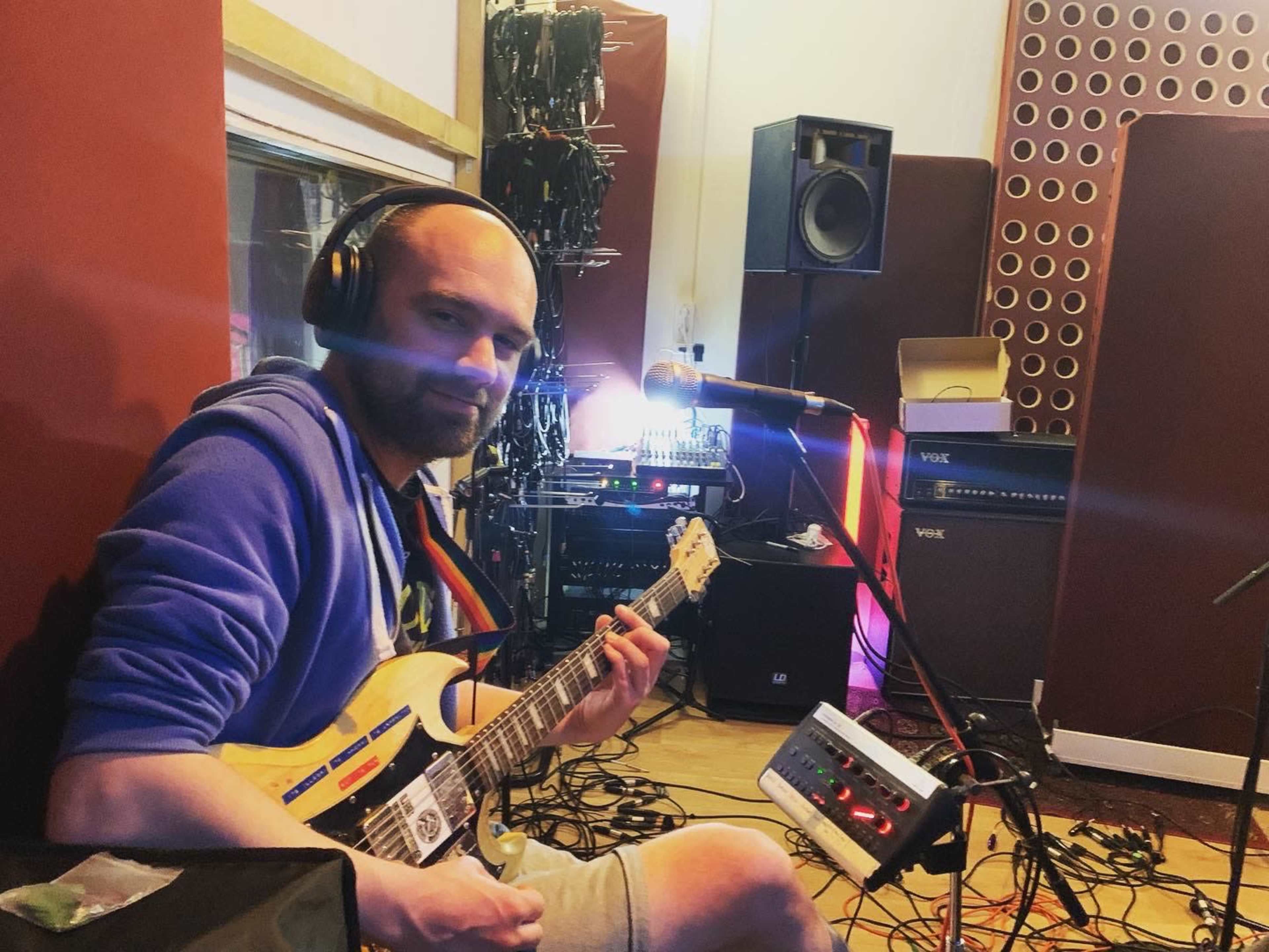 A man in a blue hoodie plays an electric guitar in a recording studio surrounded by musical equipment and cables.
