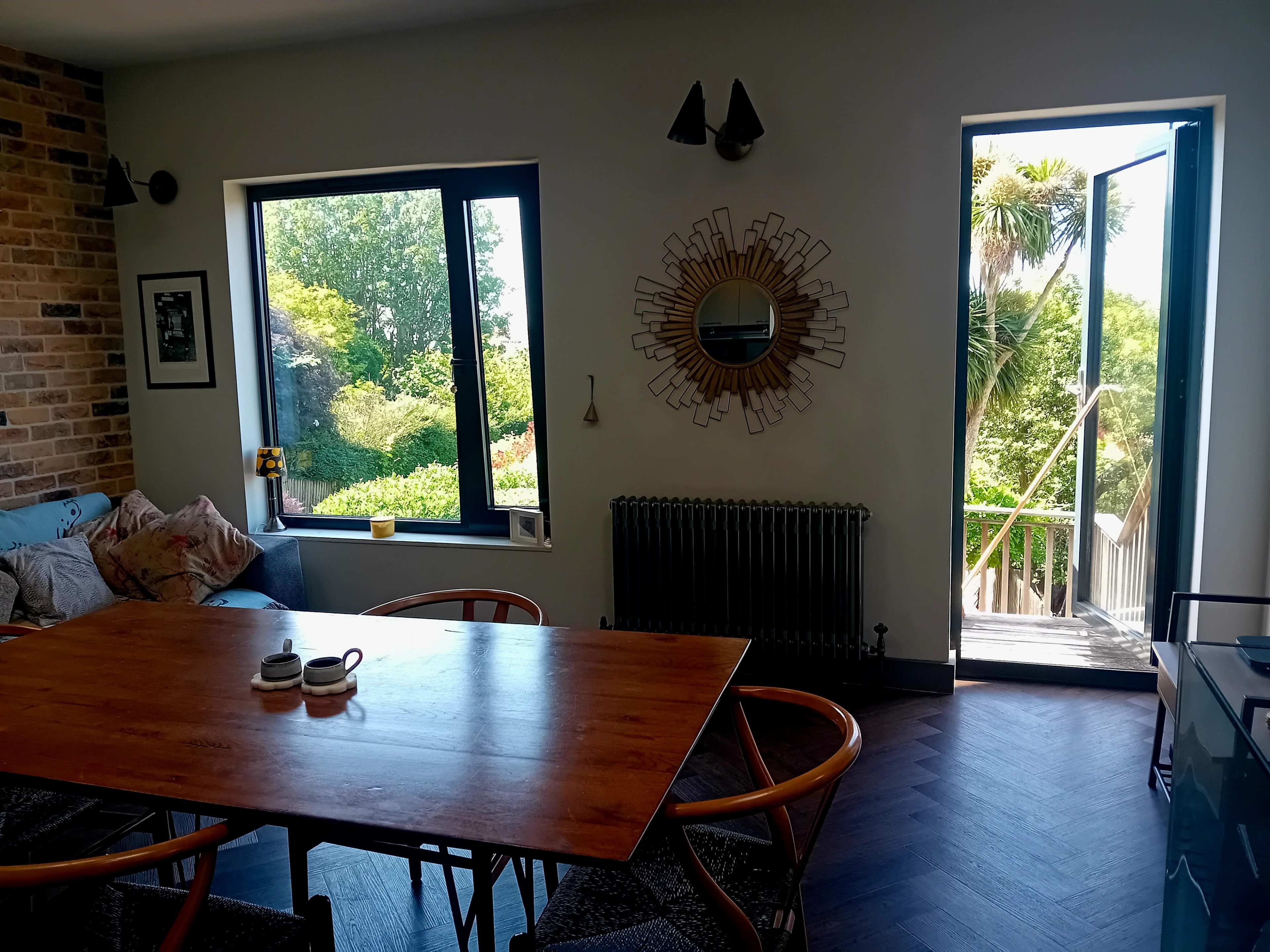 A dining area features a wooden table surrounded by chairs, large windows overlooking a garden, and a doorway leading to a balcony.