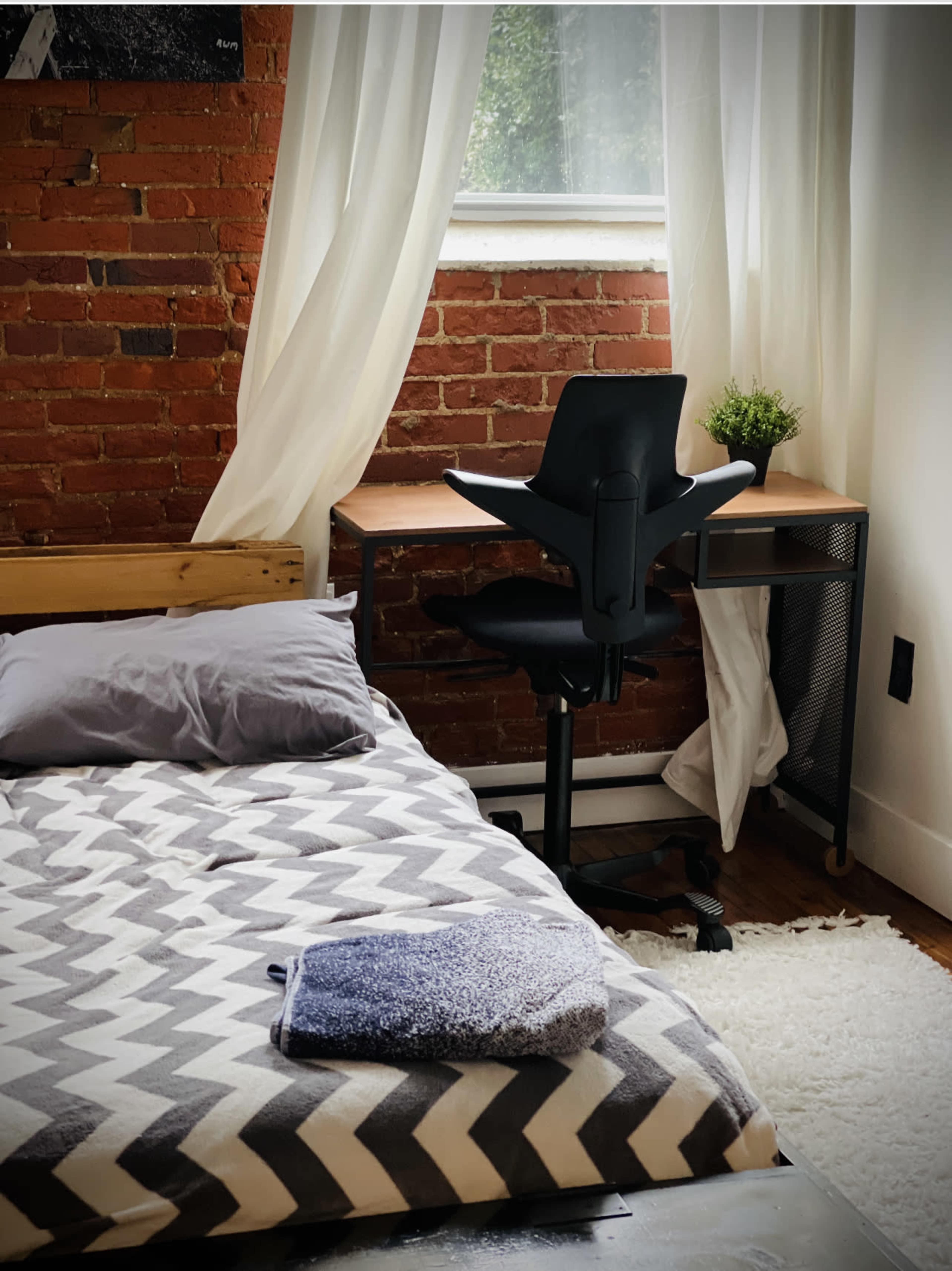 A neatly arranged bedroom features a bed with a patterned blanket and a desk with an office chair near a window draped with sheer curtains.