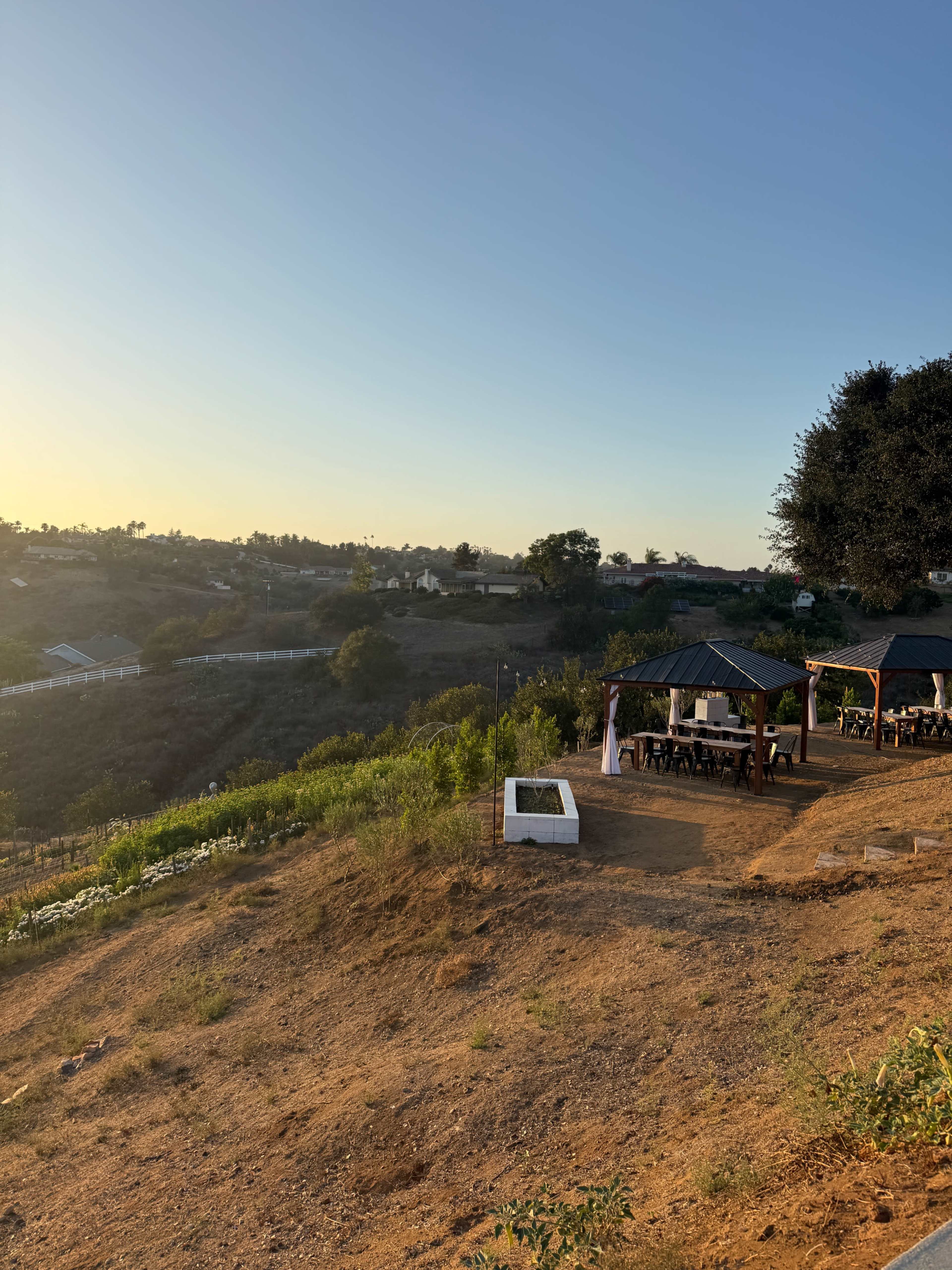 The image shows a hillside landscape with two wooden gazebos overlooking a valley during sunset.