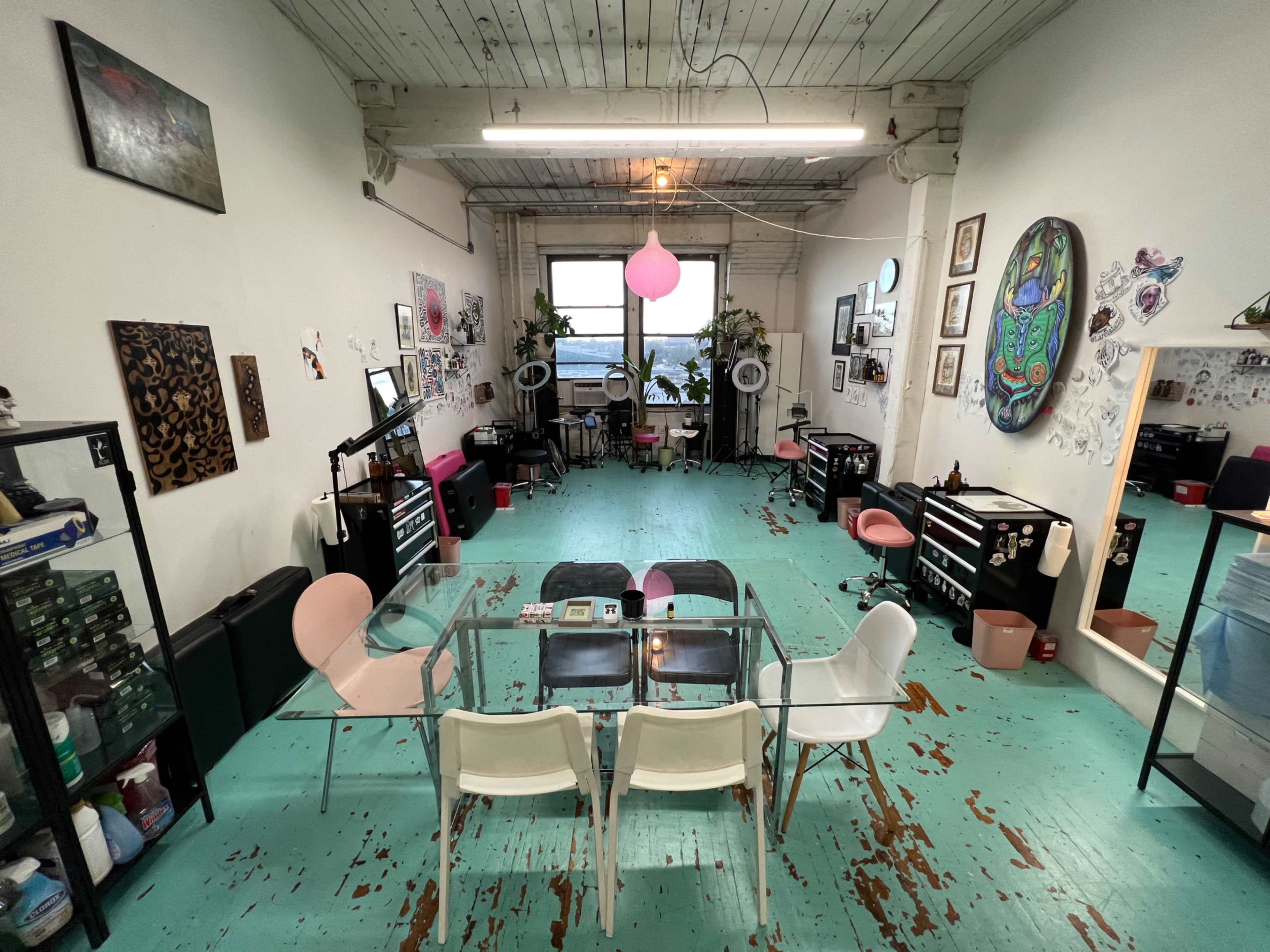 A spacious tattoo studio with green wooden floors, several workstations, and a glass table surrounded by chairs.