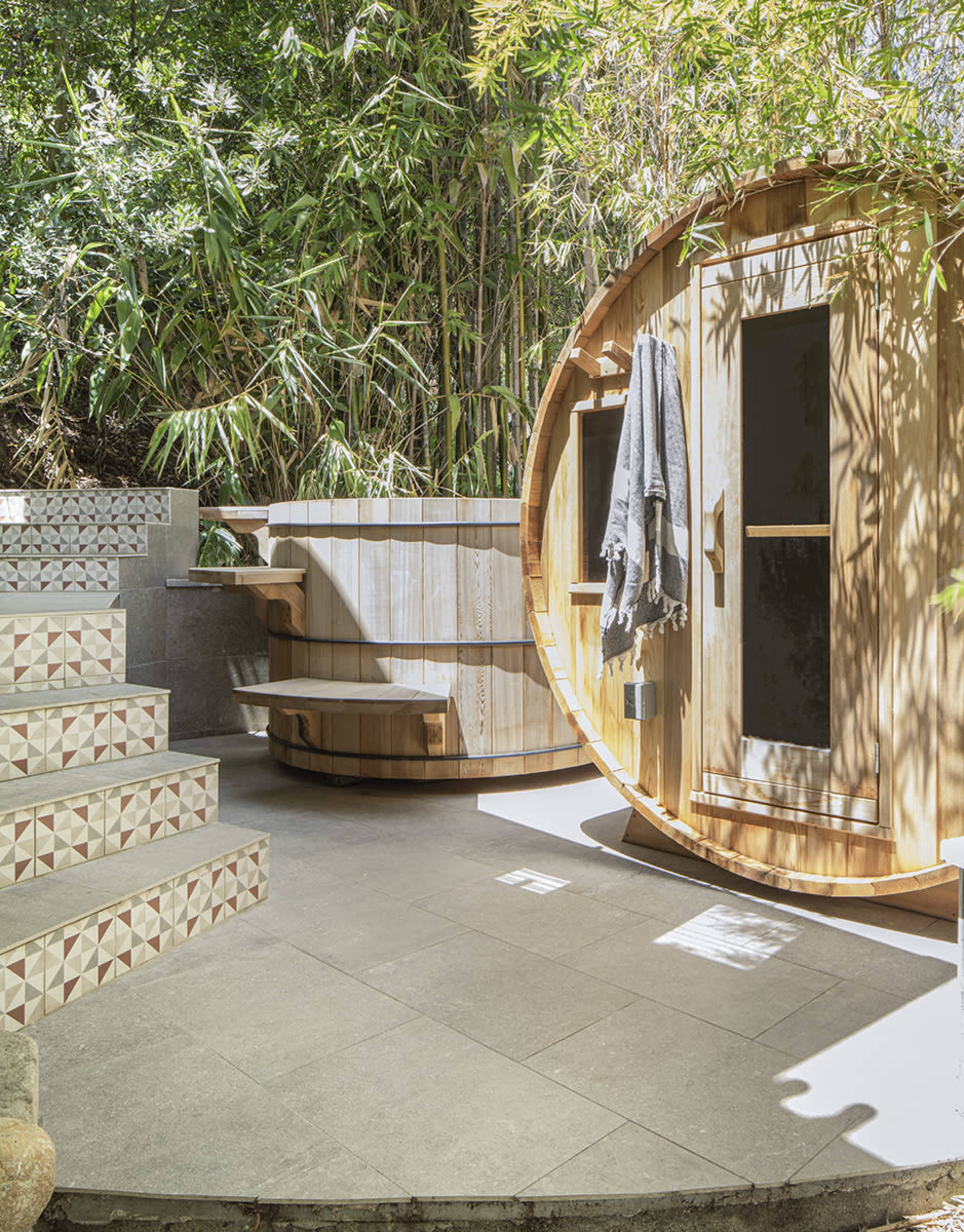The scene features a circular wooden sauna adjacent to a round hot tub, surrounded by bamboo and stone steps.