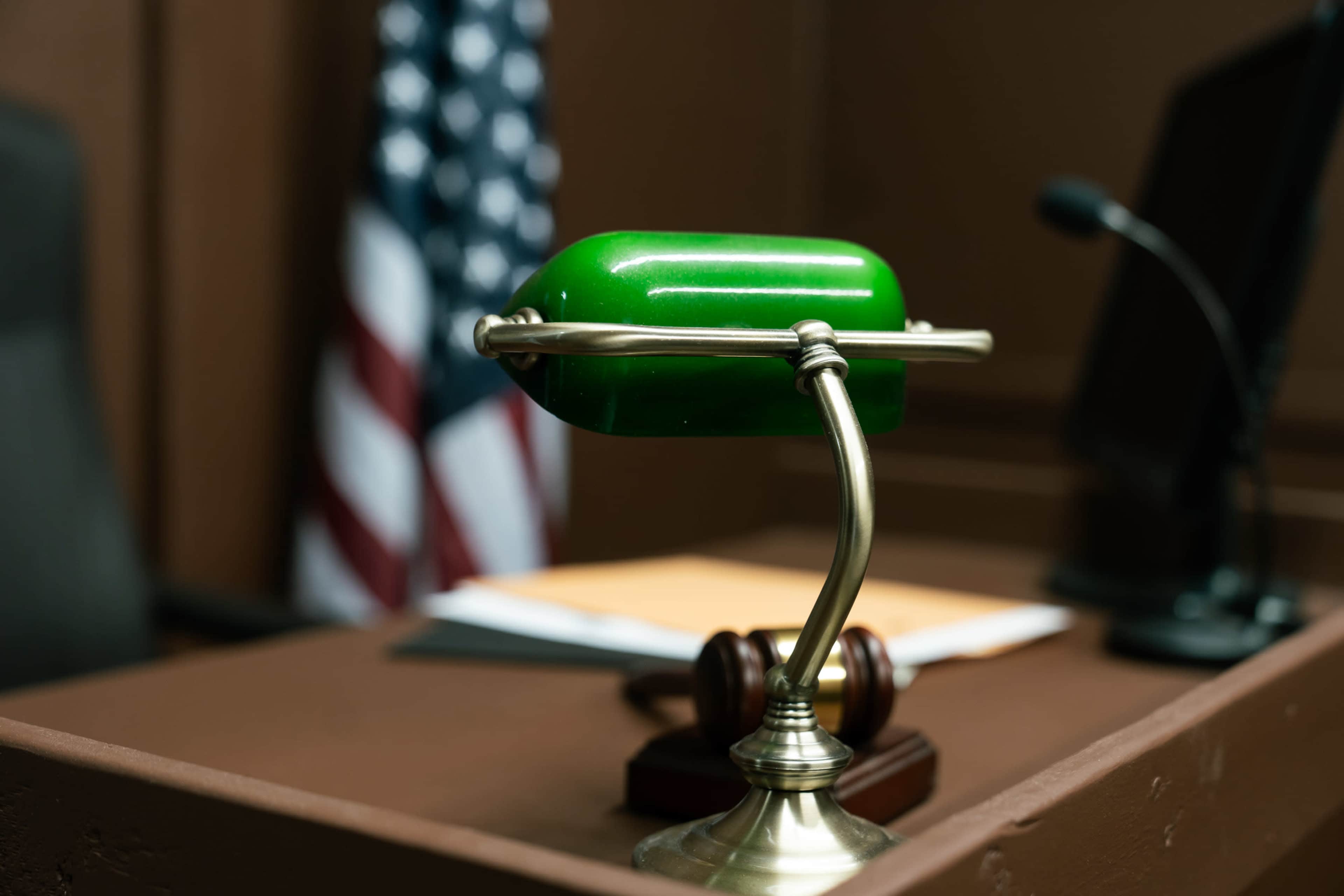 A green desk lamp is positioned on a wooden court table with an American flag in the background.