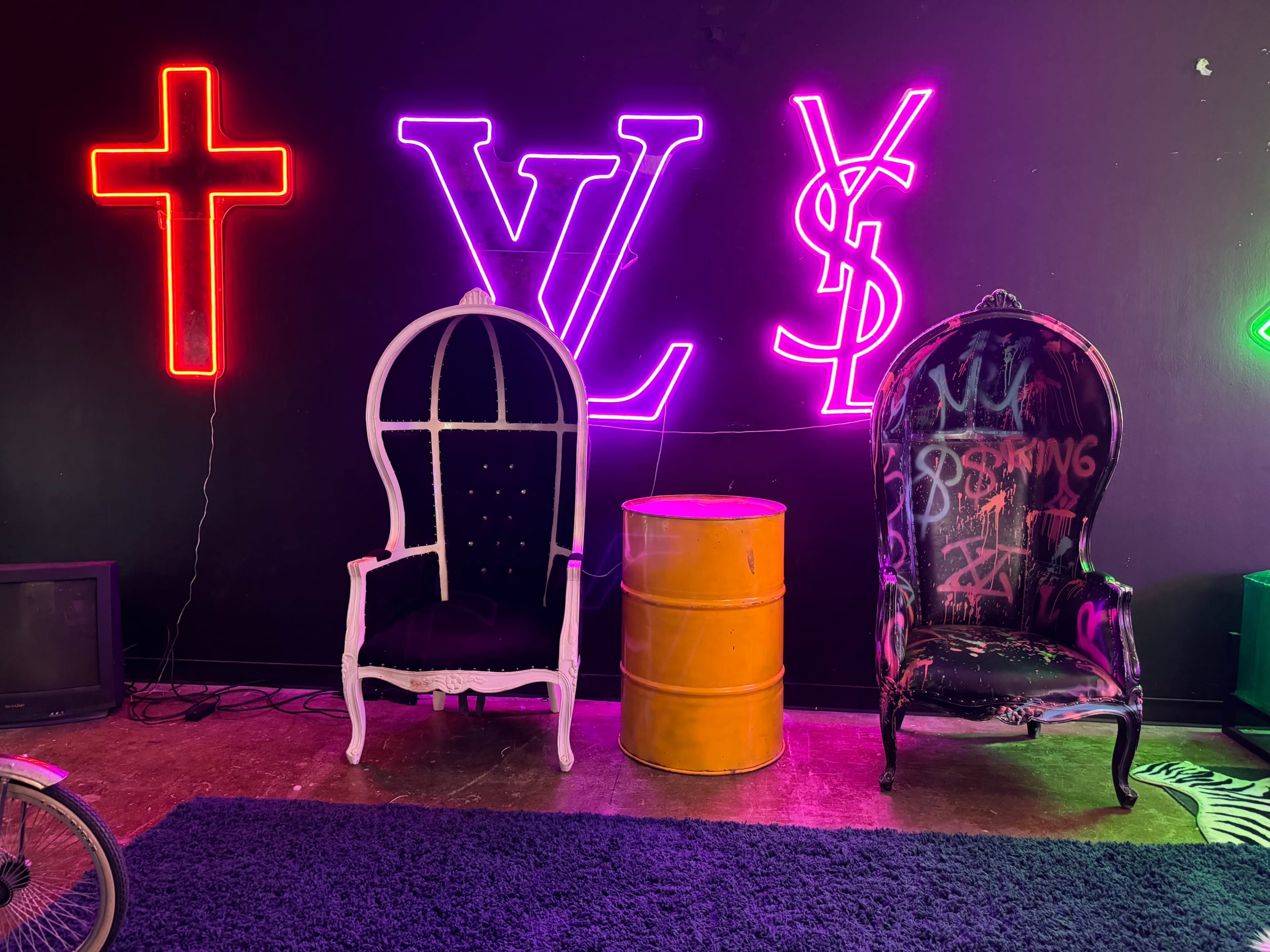 The scene features two distinct chairs beside a yellow barrel, with neon signs displaying symbols and letters on the black wall behind them.