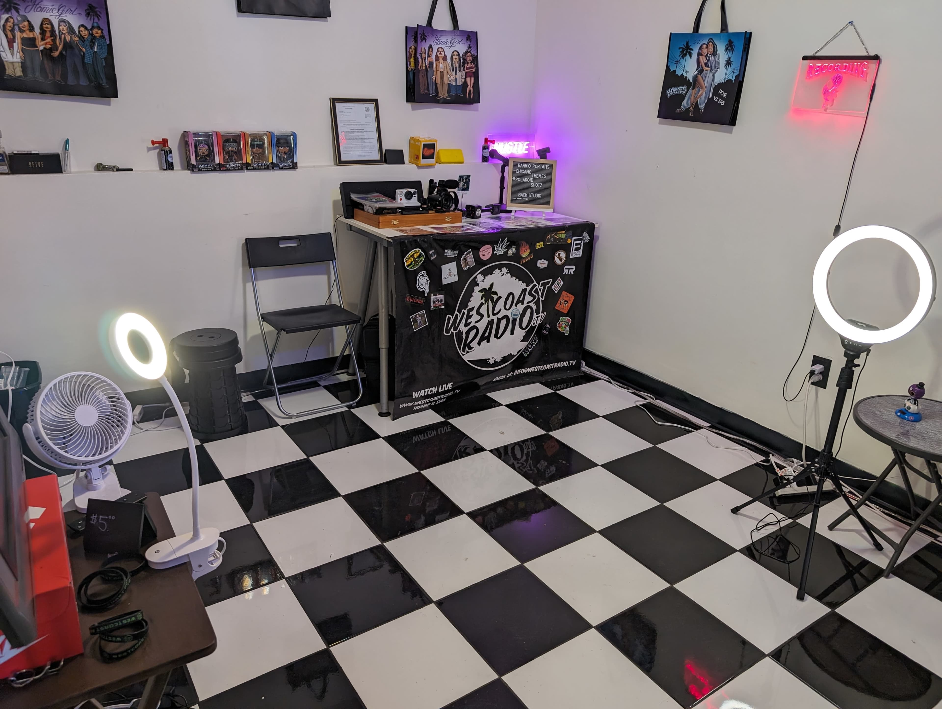 A corner of a studio space with a checkered floor, a desk featuring a computer and various equipment, and lighting equipment on either side.