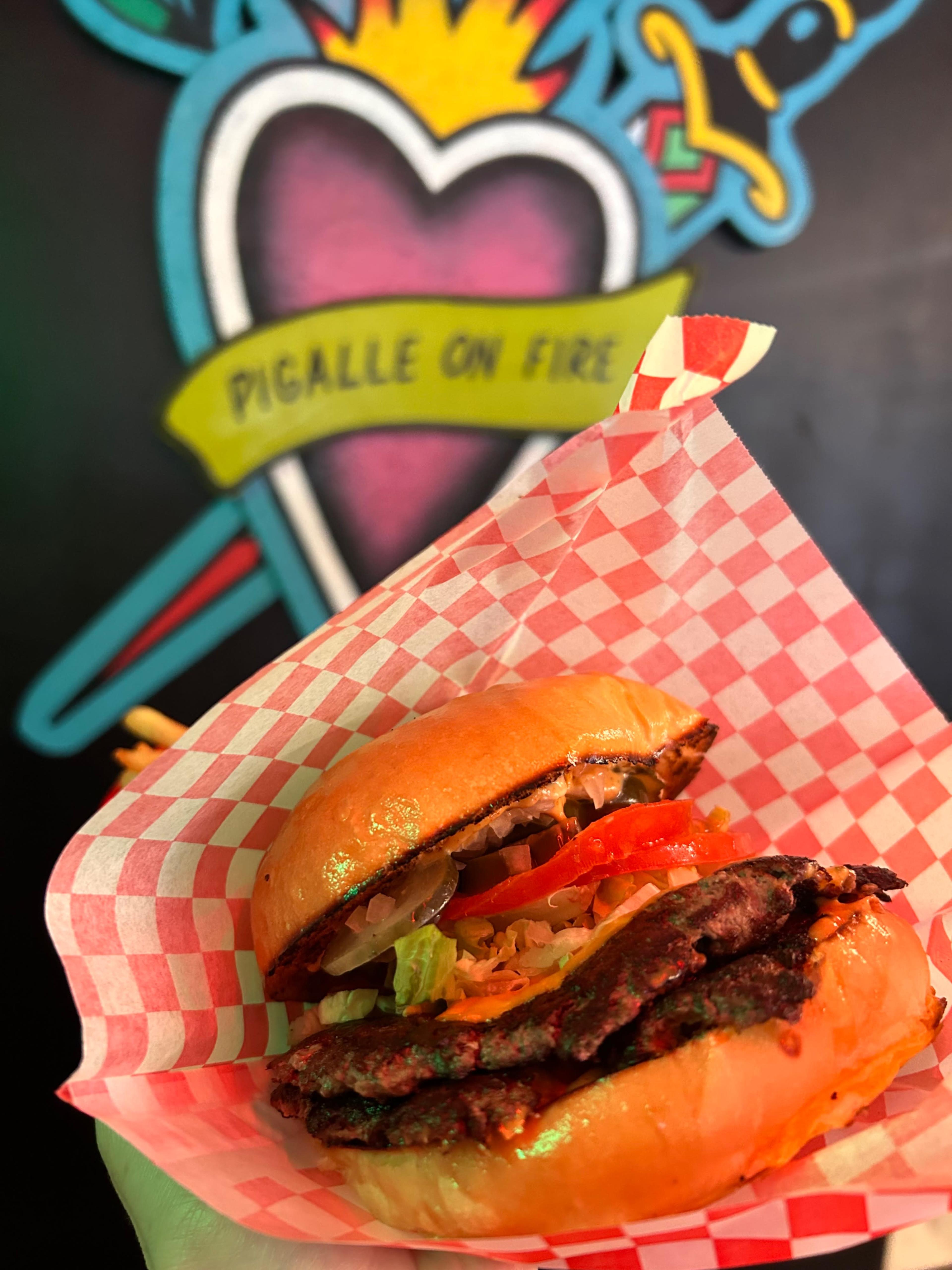 A hand holds a hamburger with toppings, positioned in front of a colorful mural that features a heart and the words "Pigalle on Fire."