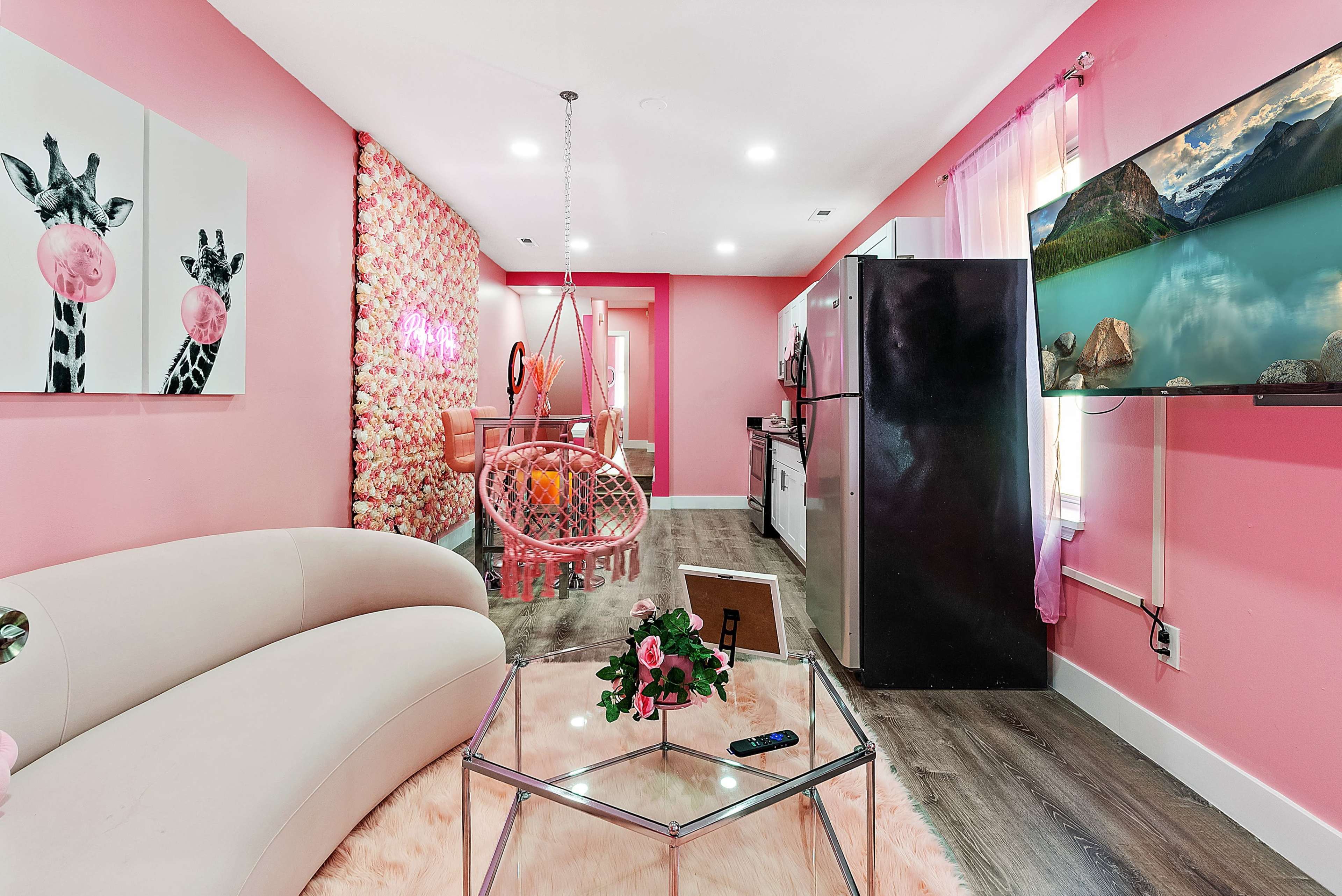 A pink-themed living space featuring a curved sofa, a glass coffee table, a hanging chair, and a kitchen area with black appliances.
