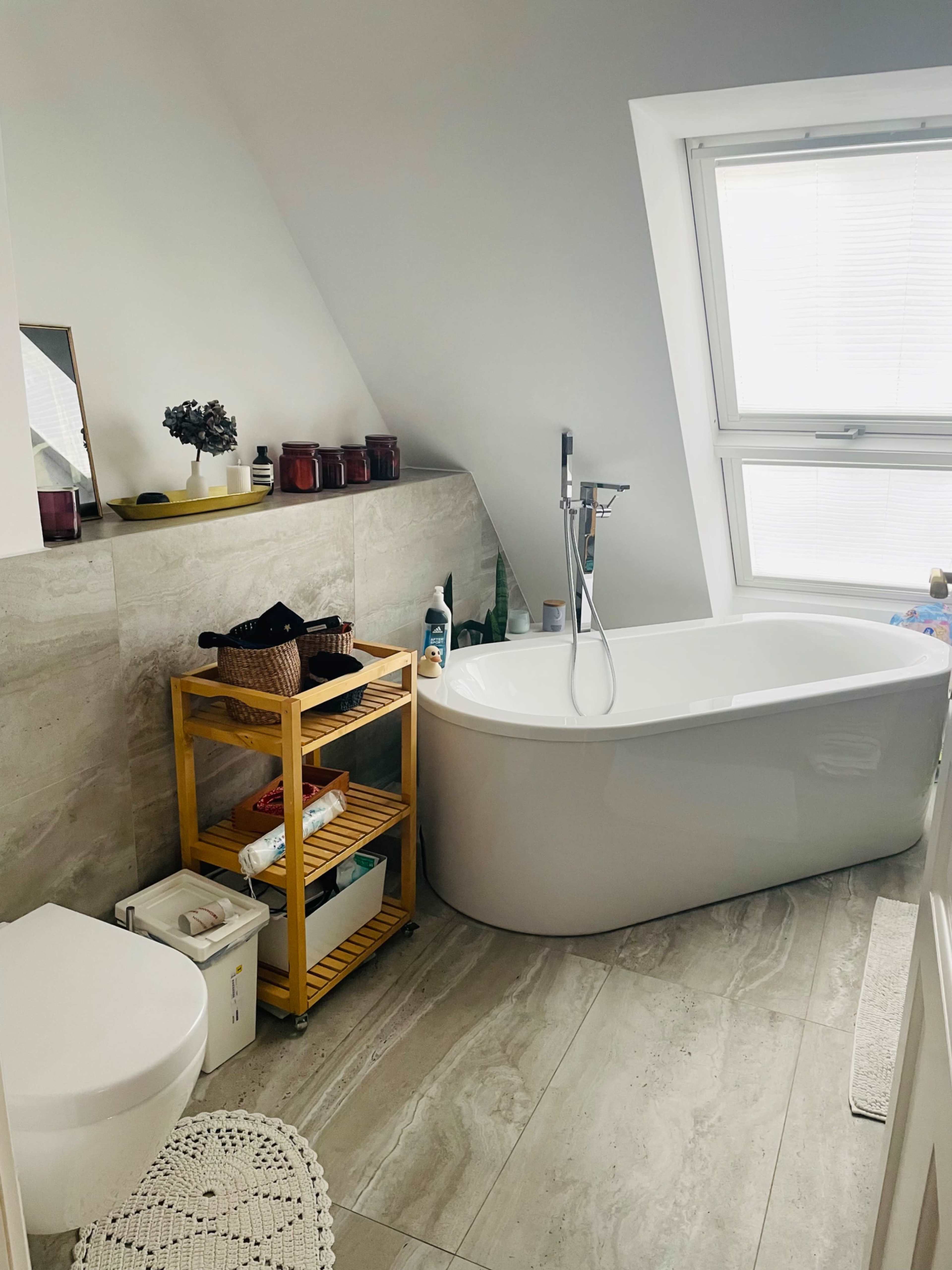 The bathroom features a freestanding bathtub, a modern sink area, a wooden storage cart, and a large window allowing natural light.