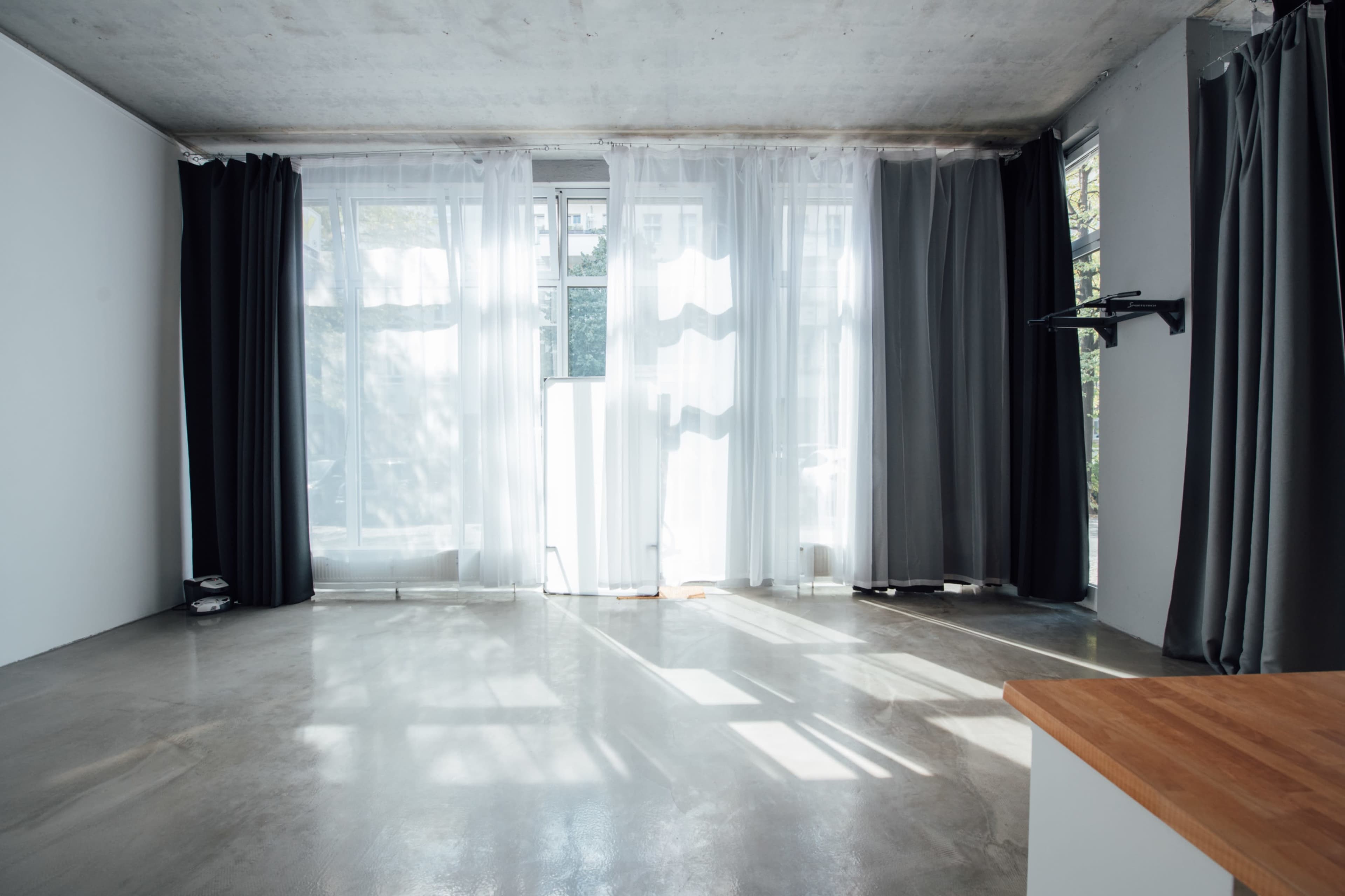 The image shows a minimalist interior space with large windows draped in sheer and dark curtains, allowing sunlight to cast patterns on the polished concrete floor.