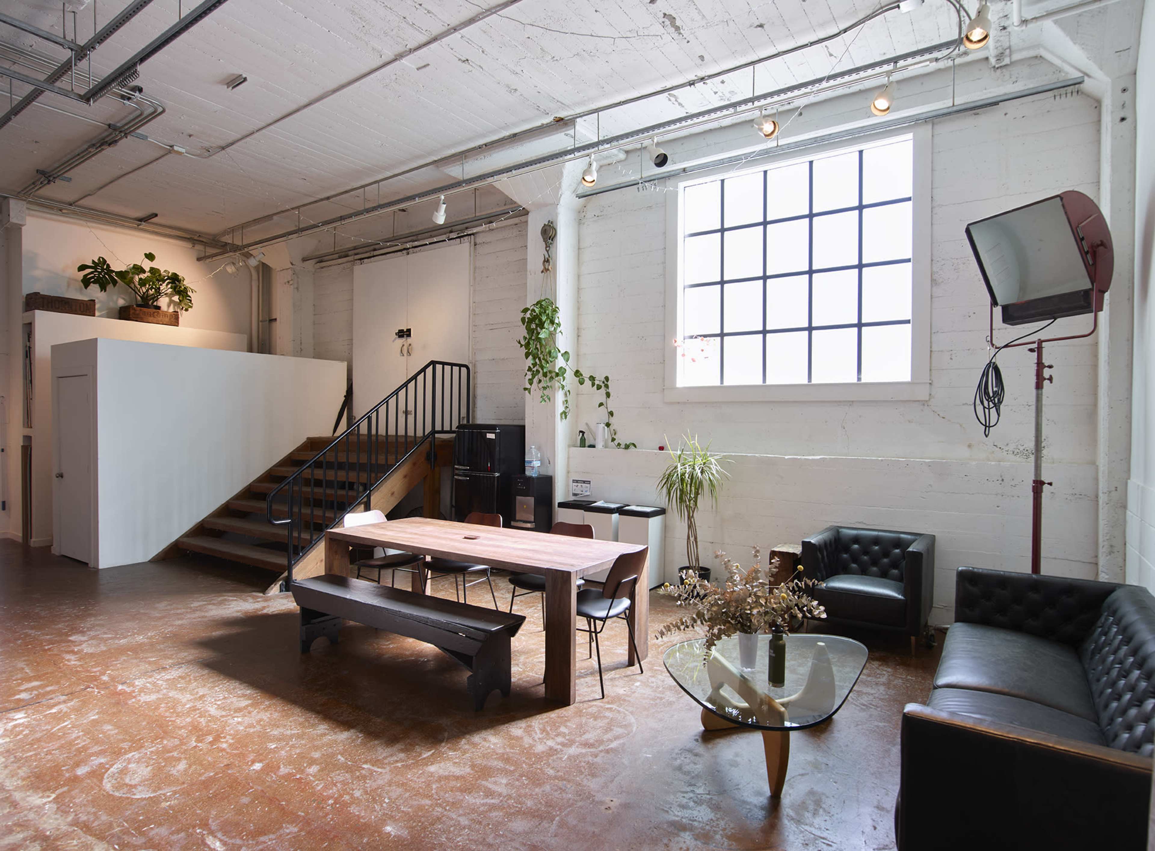 The image shows a spacious industrial-style room with wooden floors, a large table in the center, a seating area with a black leather couch, and a staircase leading to a higher level.