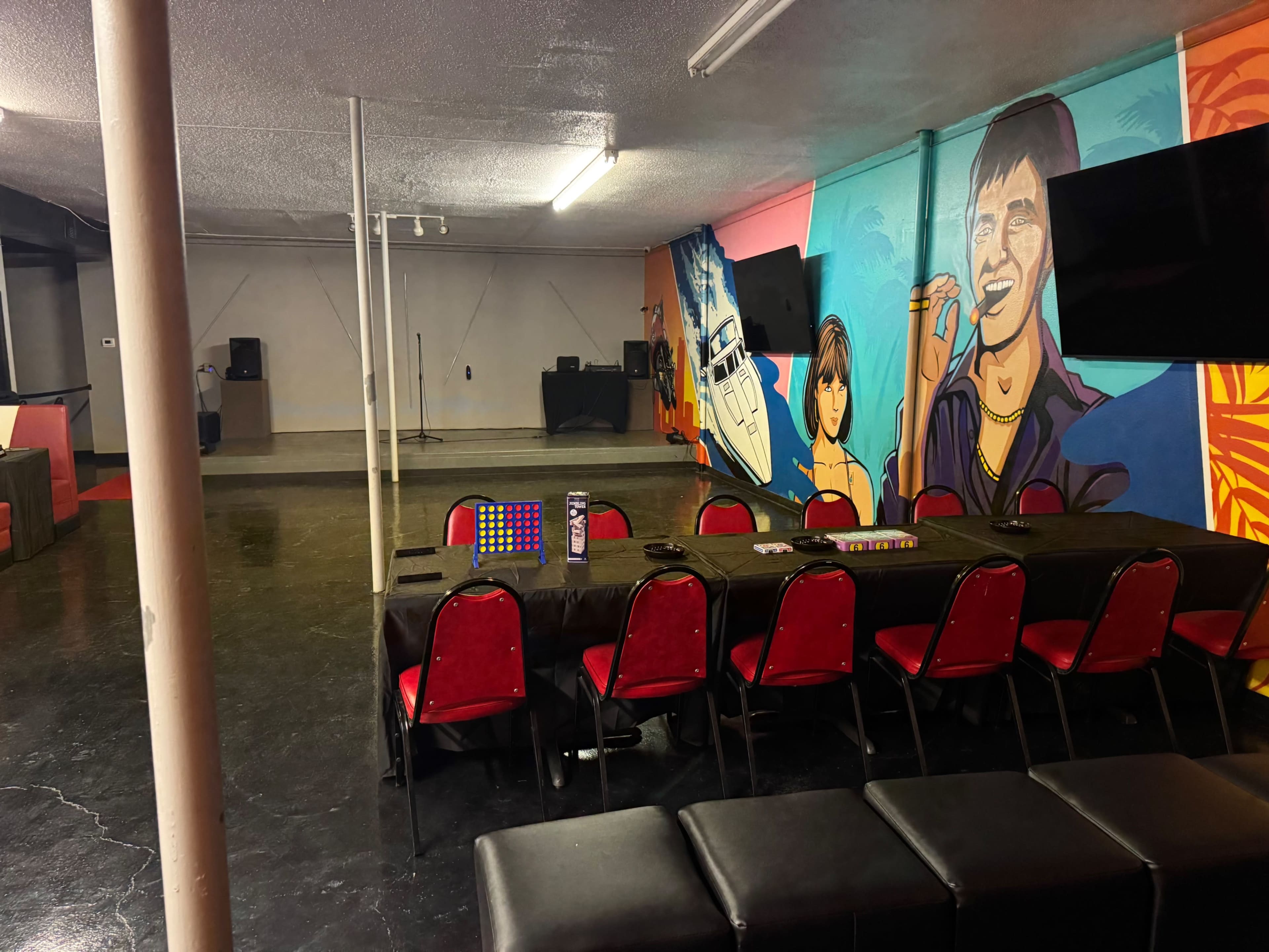 The image shows an indoor space featuring a long table with red chairs, a large mural on the wall, and electronic equipment in the background.