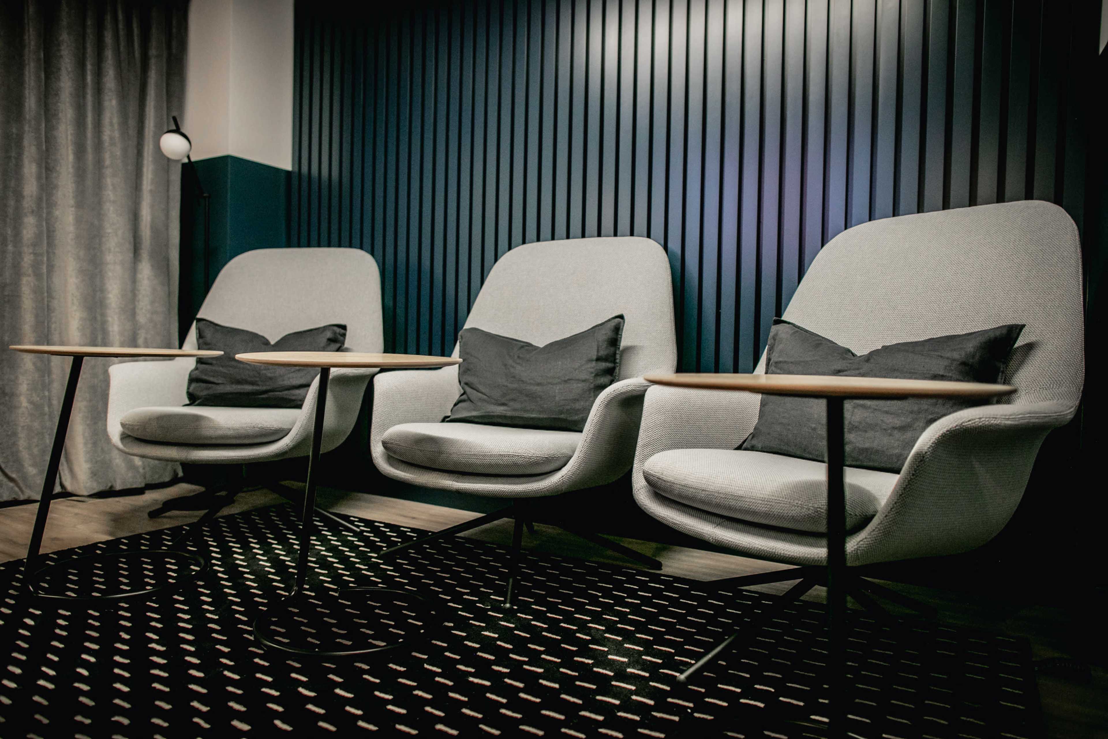 Three modern gray chairs with cushions, paired with small round wooden tables, arranged in a stylish indoor space with a textured dark wall.