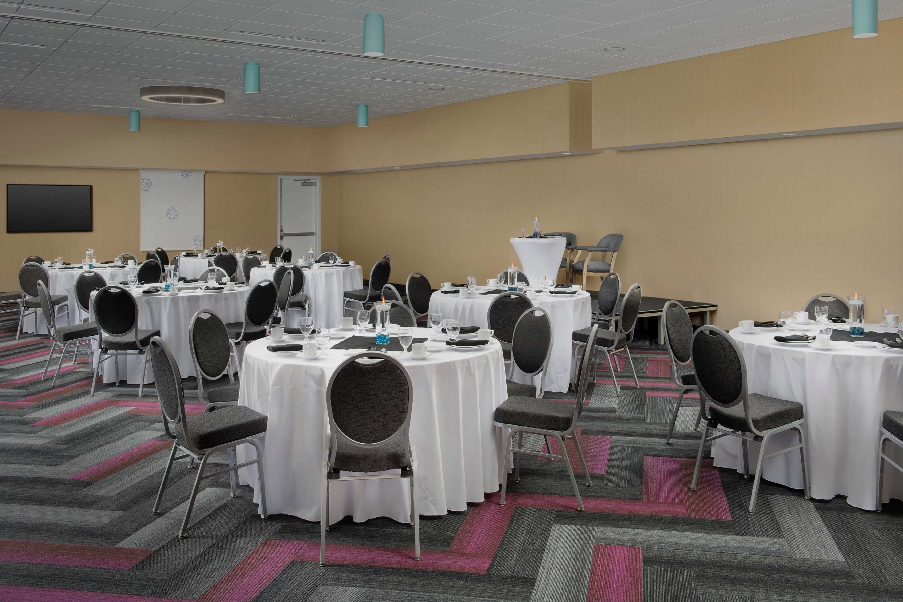 The image shows a banquet hall arranged with multiple round tables covered in white tablecloths, each set with dinnerware and black chairs, and featuring a modern carpet design in shades of gray and pink.