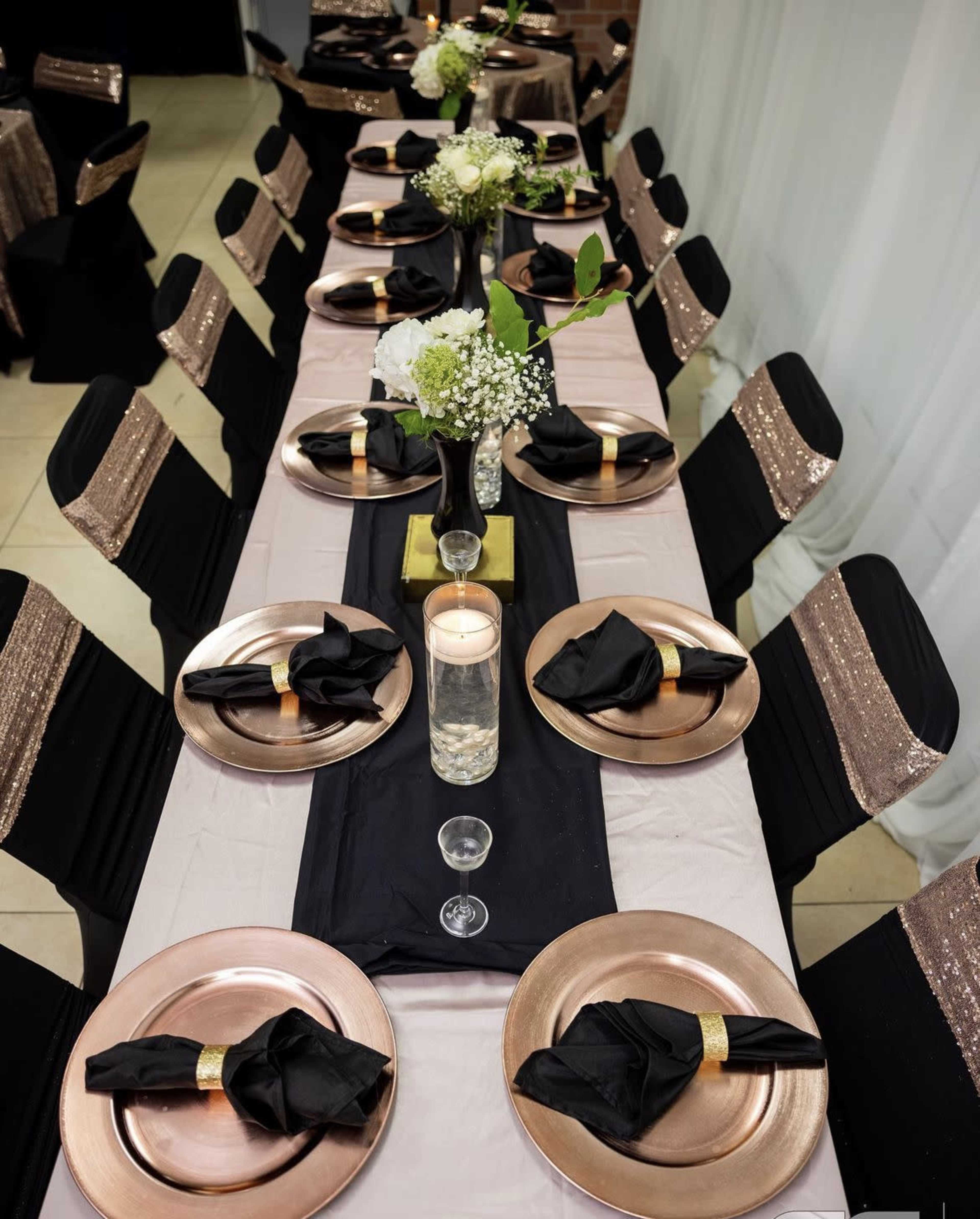 A long dining table is set with gold-rimmed plates, black napkins, and floral centerpieces on a black runner, surrounded by chairs with sparkling black covers.