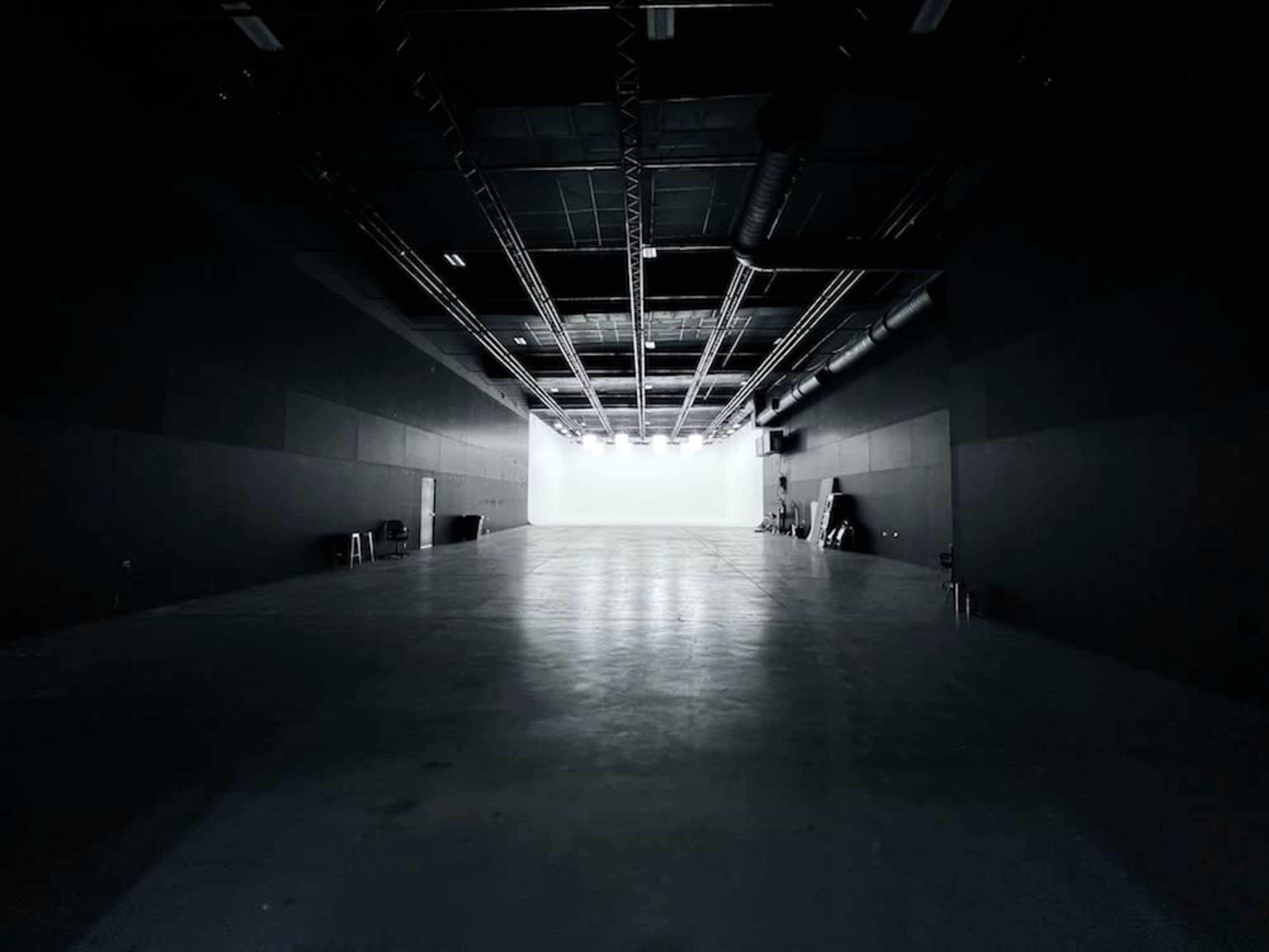 A large, empty studio space with dark walls and bright lights at the far end illuminating a white backdrop.