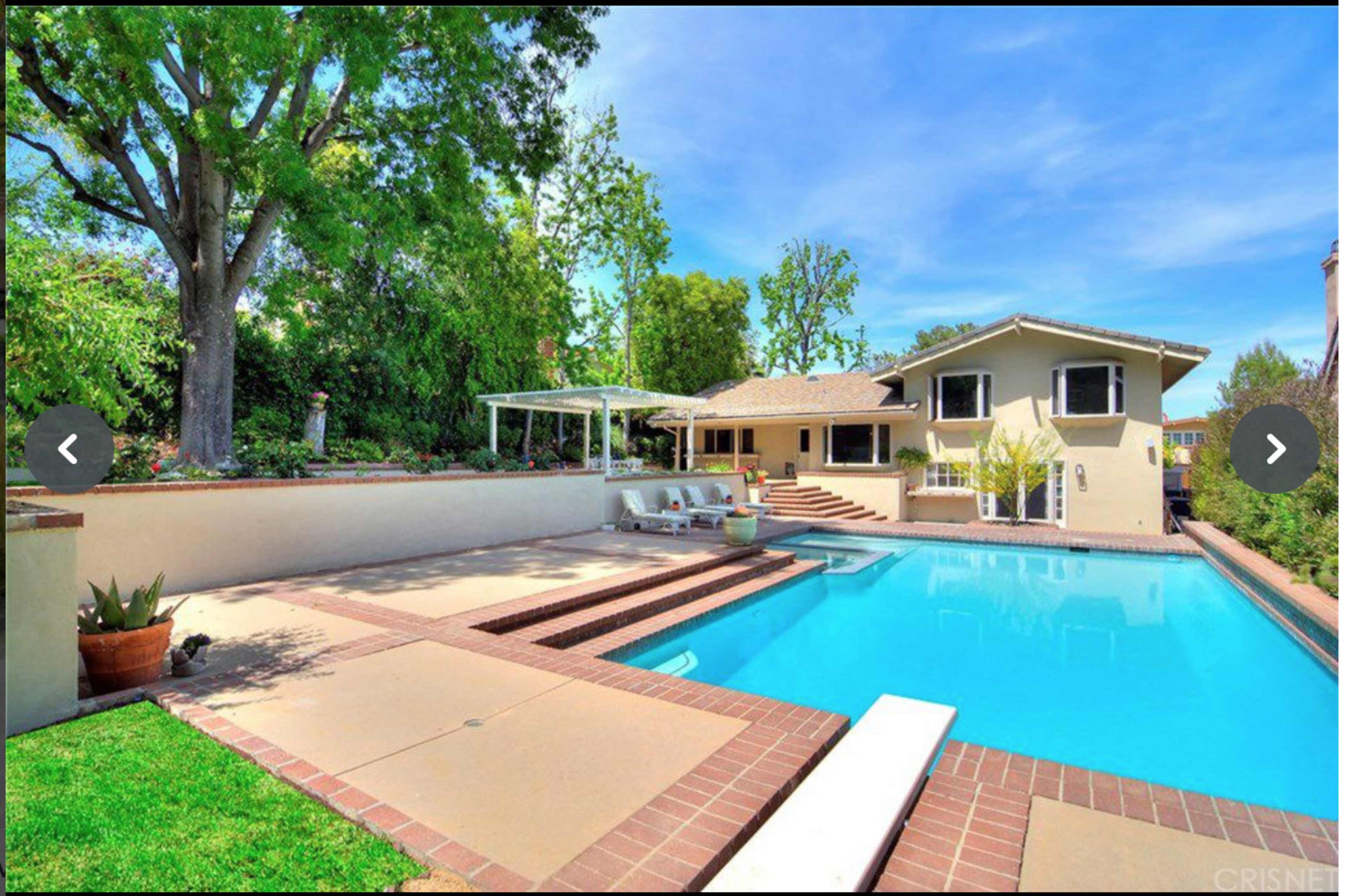 A spacious backyard featuring a swimming pool, lounge area, and a house with a patio surrounded by trees and greenery.