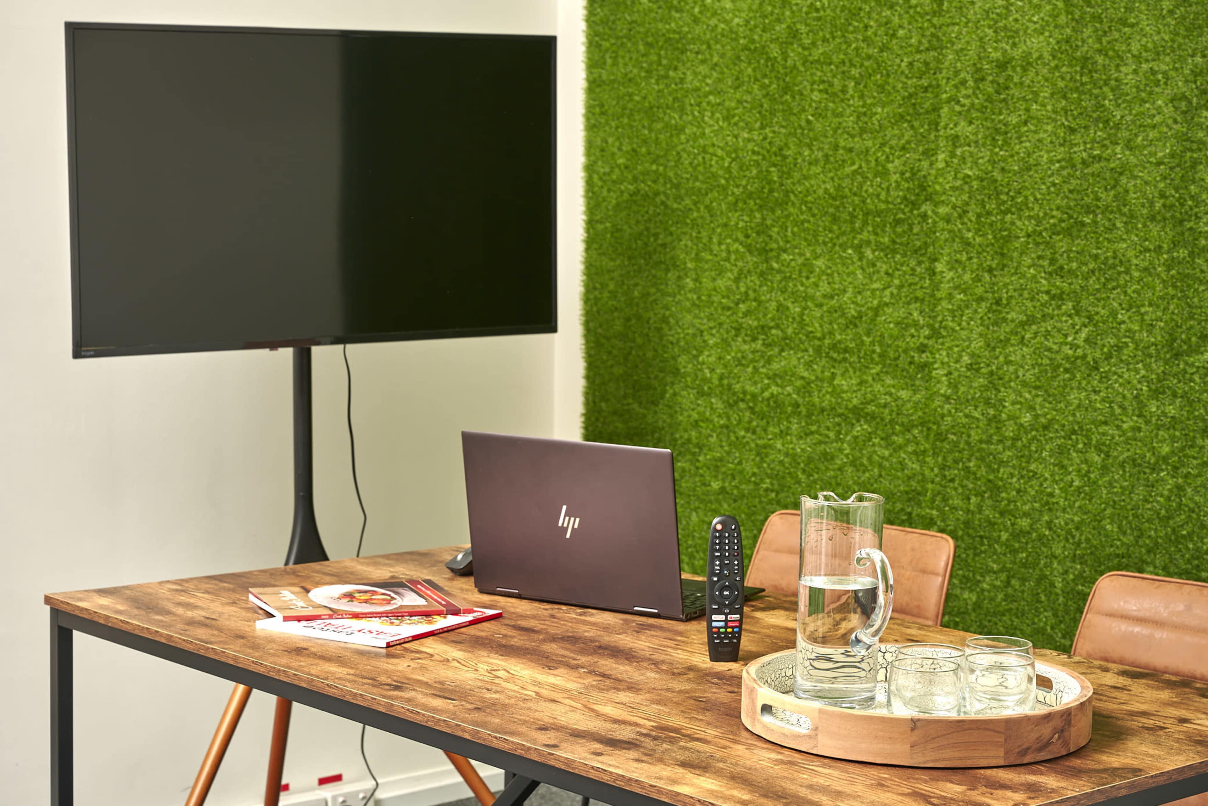 A wooden table in an office setting features a laptop, a remote control, a pitcher of water, and glasses, with a large TV mounted on a wall covered in green foliage.