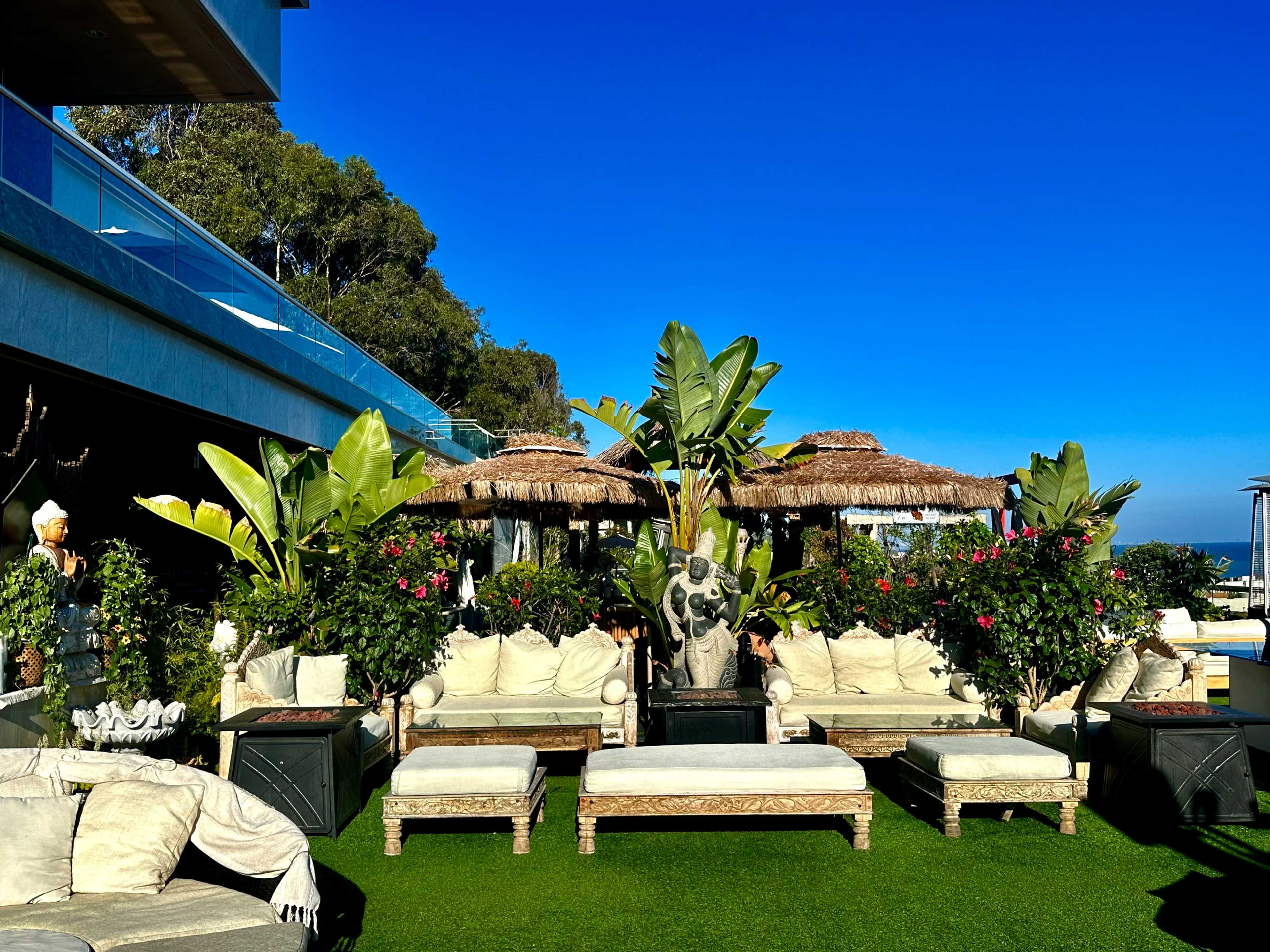 A stylish outdoor lounge area with wooden seating, thatched umbrellas, and lush greenery under a clear blue sky.